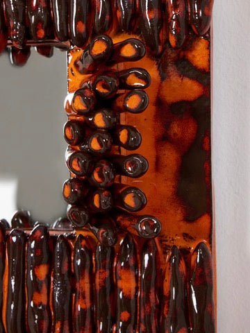 A close-up of the 'Dedo' Ceramic Mirror by Project 213A, showcasing its glossy, textured surface with dark red, orange, and black hues. It features tube-like protrusions around a rectangular shape that reflect light against a partial view of the background wall.