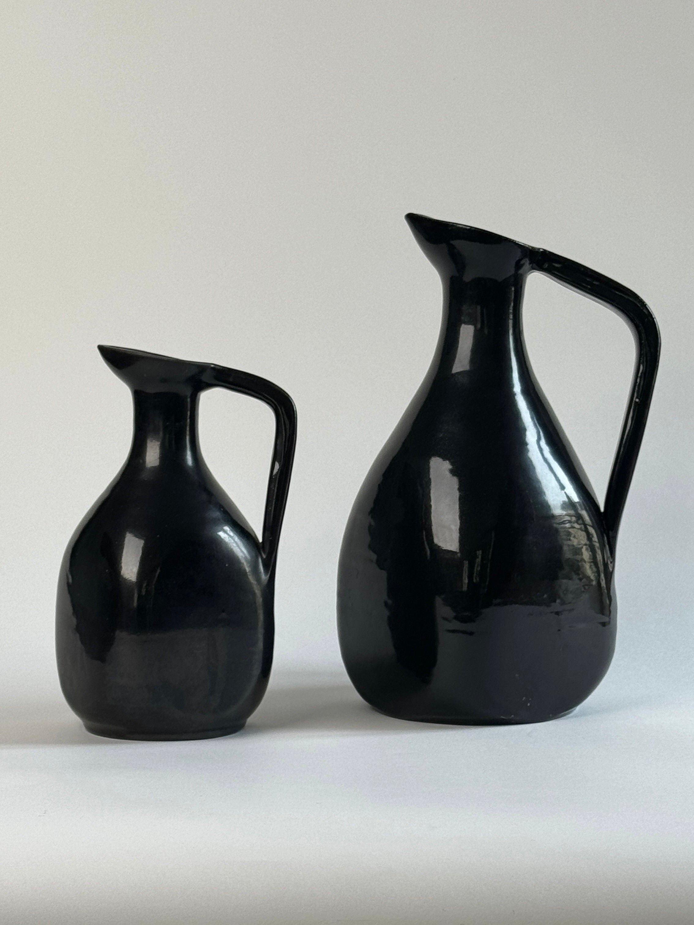 A photograph of the Pair of Accolay Ceramic Carafes by Figuresdesign shows two black ceramic pitchers with handles, one smaller and one larger, placed side by side against a plain light-colored background. The glossy finish on these vintage French pottery pieces reflects the light, highlighting their elegant design.