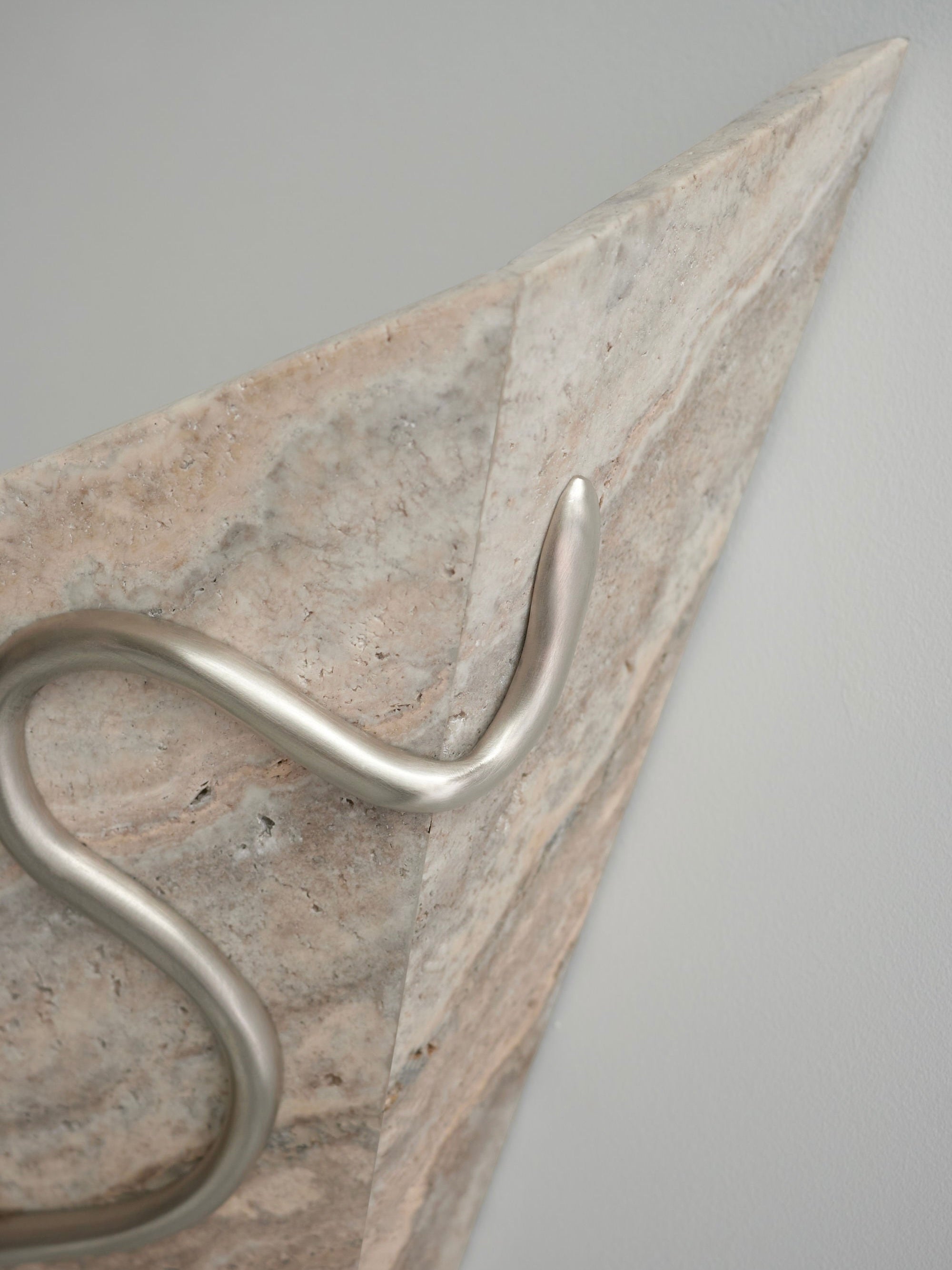 A close-up image of Mariza Galani's Therapeutic Wall Light 3, a triangular wall sconce with a marble texture and sleek, metallic curved support. This therapeutic fixture features a blend of light beige and off-white hues and is mounted against a plain white wall.