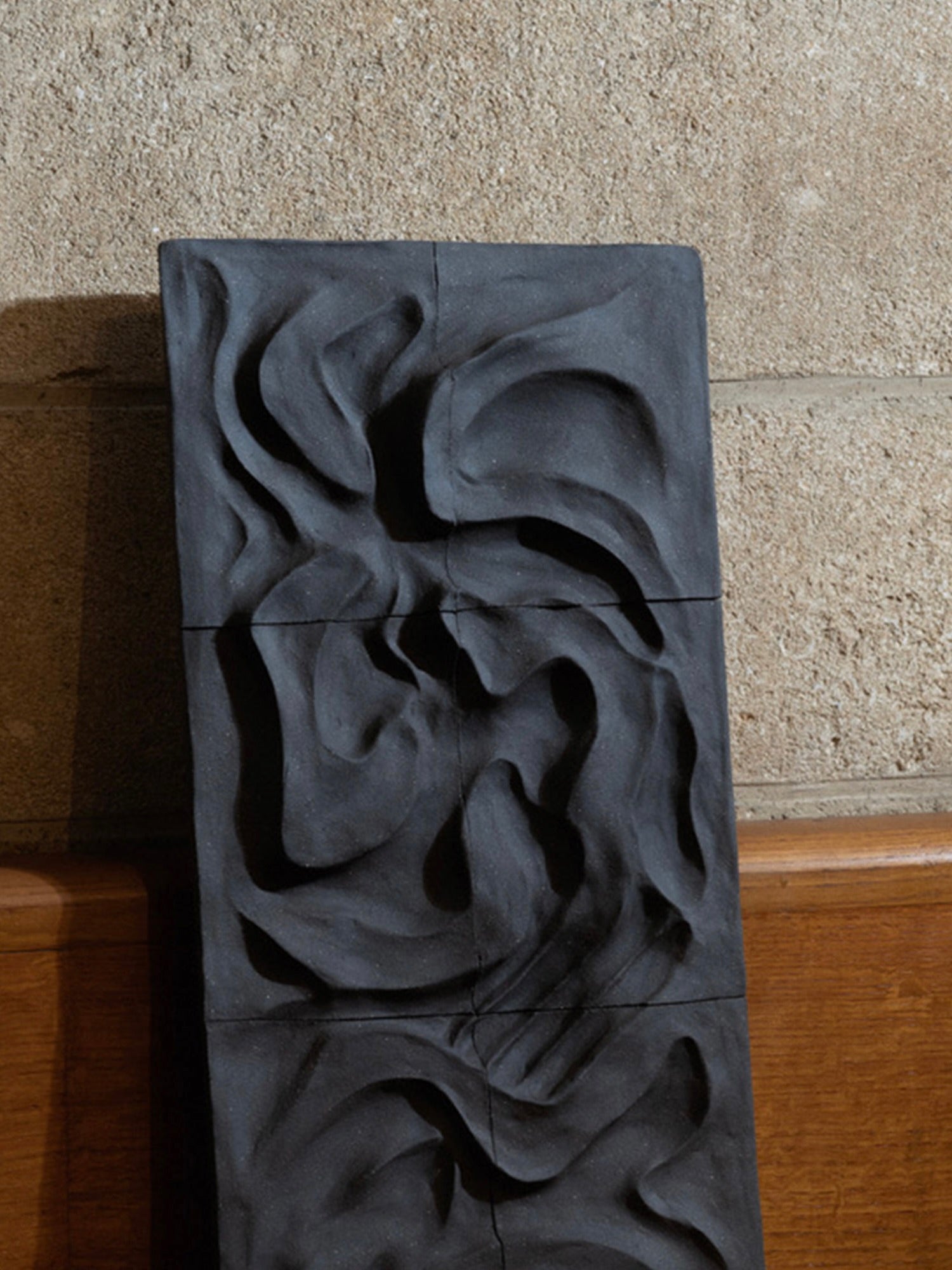 The Kilauea Lava wall sculpture by Franck Scala, mounted on a light stone wall above a wooden surface, features dark, abstract reliefs with swirling, wave-like patterns that evoke movement and texture similar to intricate ceramic bas-reliefs.