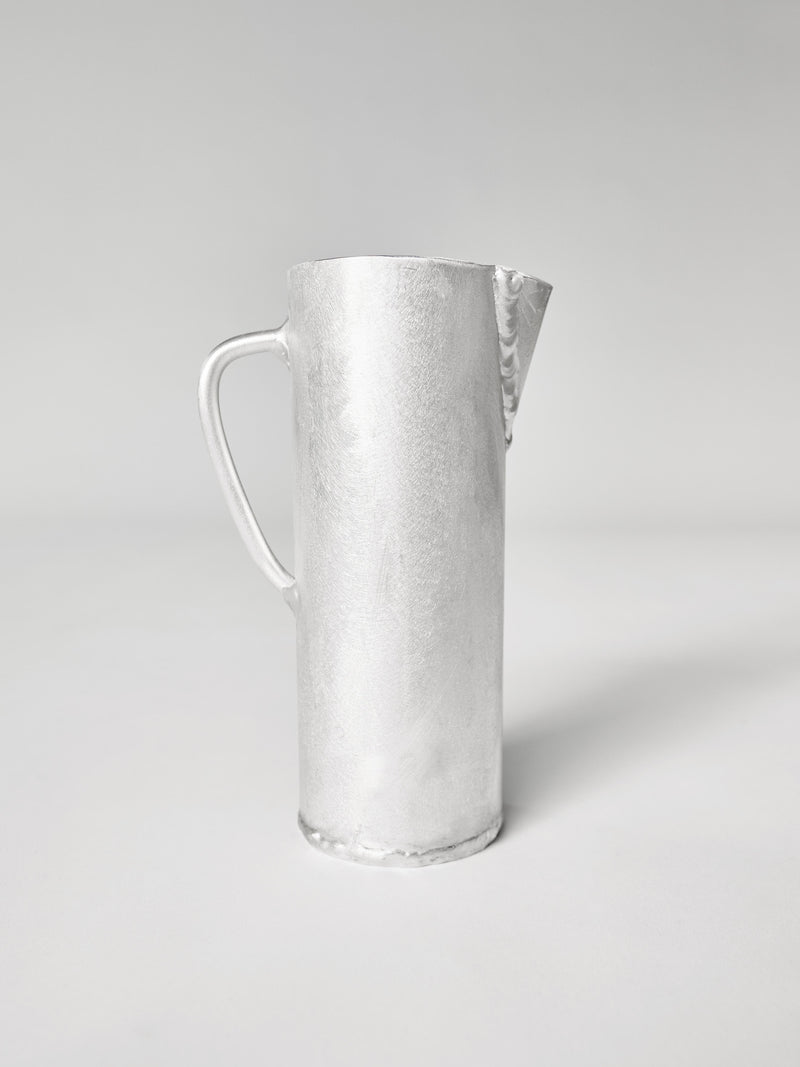 The Aluminum Jug by Six Dots Design is a silver pitcher with a minimalist design, showcasing a cylindrical body, small spout, and curved handle. Handcrafted in a North London studio, its simplicity is accentuated against a plain light gray background.