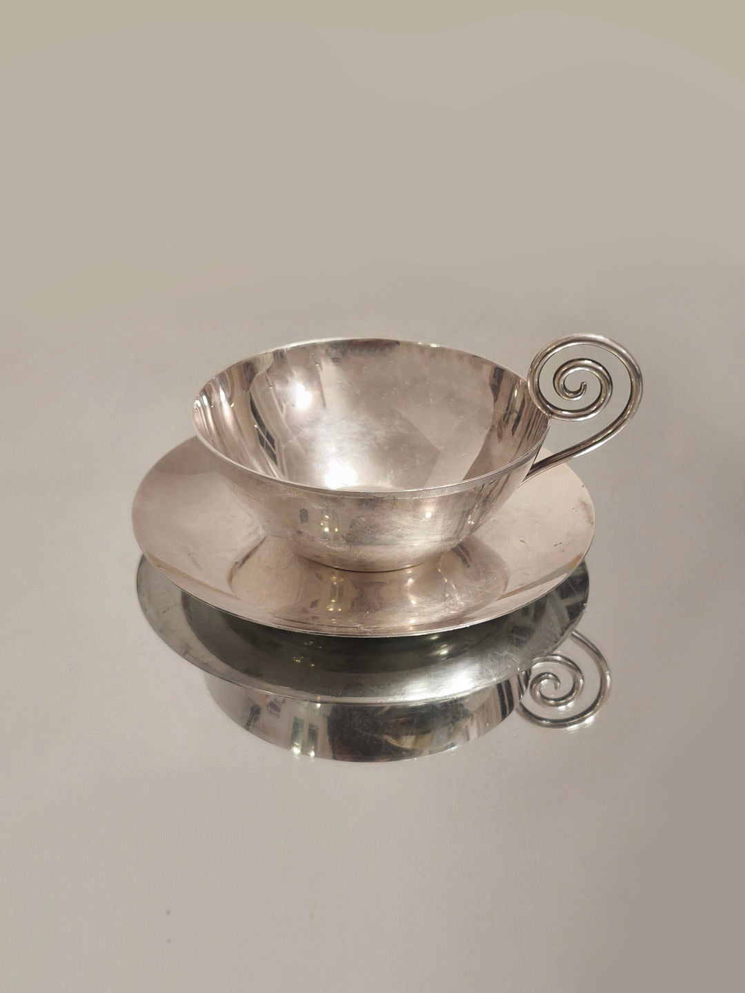 A reflective Spiral Christofle Cup from Les Objoies, featuring a decorative spiral handle, rests on a coordinating saucer. The polished surfaces mirror the table beneath it. The neutral background highlights the cup's sophisticated design and its stature as a luxury collectible.