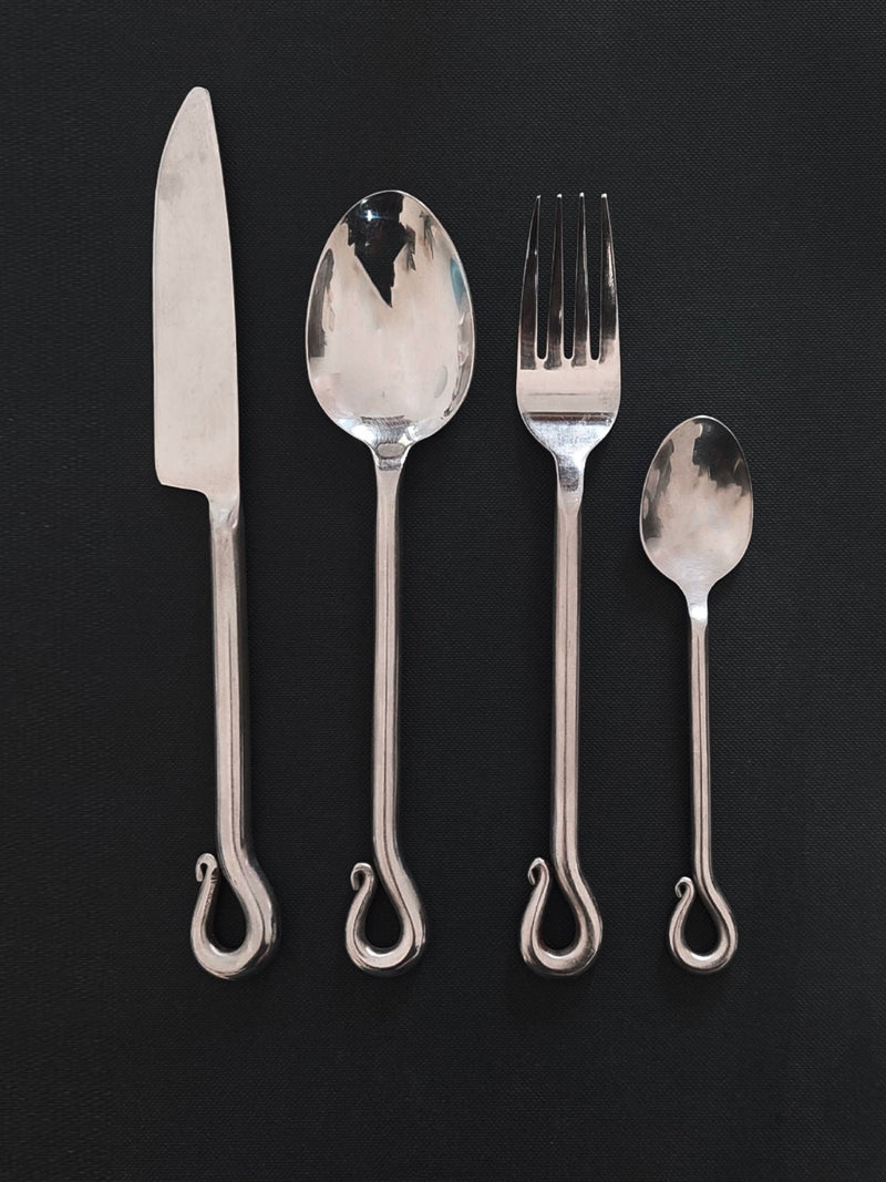 The Vintage Set of 4 Twist Cutlery by Les Objoies, reminiscent of antique utensils, is elegantly displayed against a dark background. From left to right: a knife, a tablespoon, a fork, and a teaspoon. Each piece features a distinctive curled design at the end of its handle.