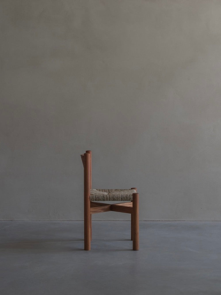 The 'Les Allues' Straw Chair by Bicci de' Medici, crafted in a minimalist style with mahogany wood and a flat woven seat, is pictured from the side against a neutral wall. The concrete floor enhances the serene atmosphere and highlights its artisanal craftsmanship.