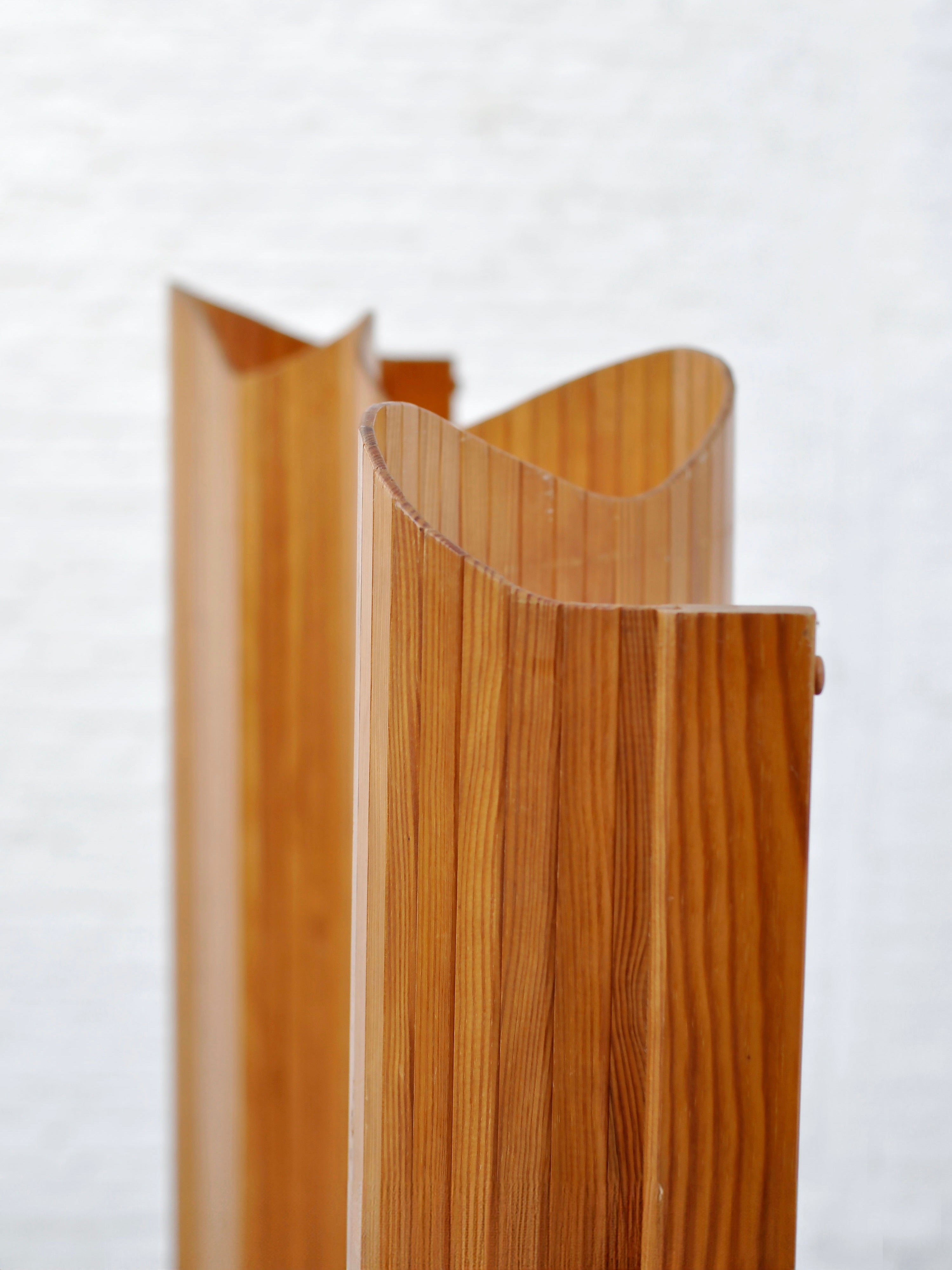 The Room Divider by Introverso, designed by Jomaine Baumann in the 1940s, showcases wooden sculptures with smooth, vertical grain against a light background. Its uniquely curved tops add artistic flair to the tambour design, reminiscent of French Art Deco.