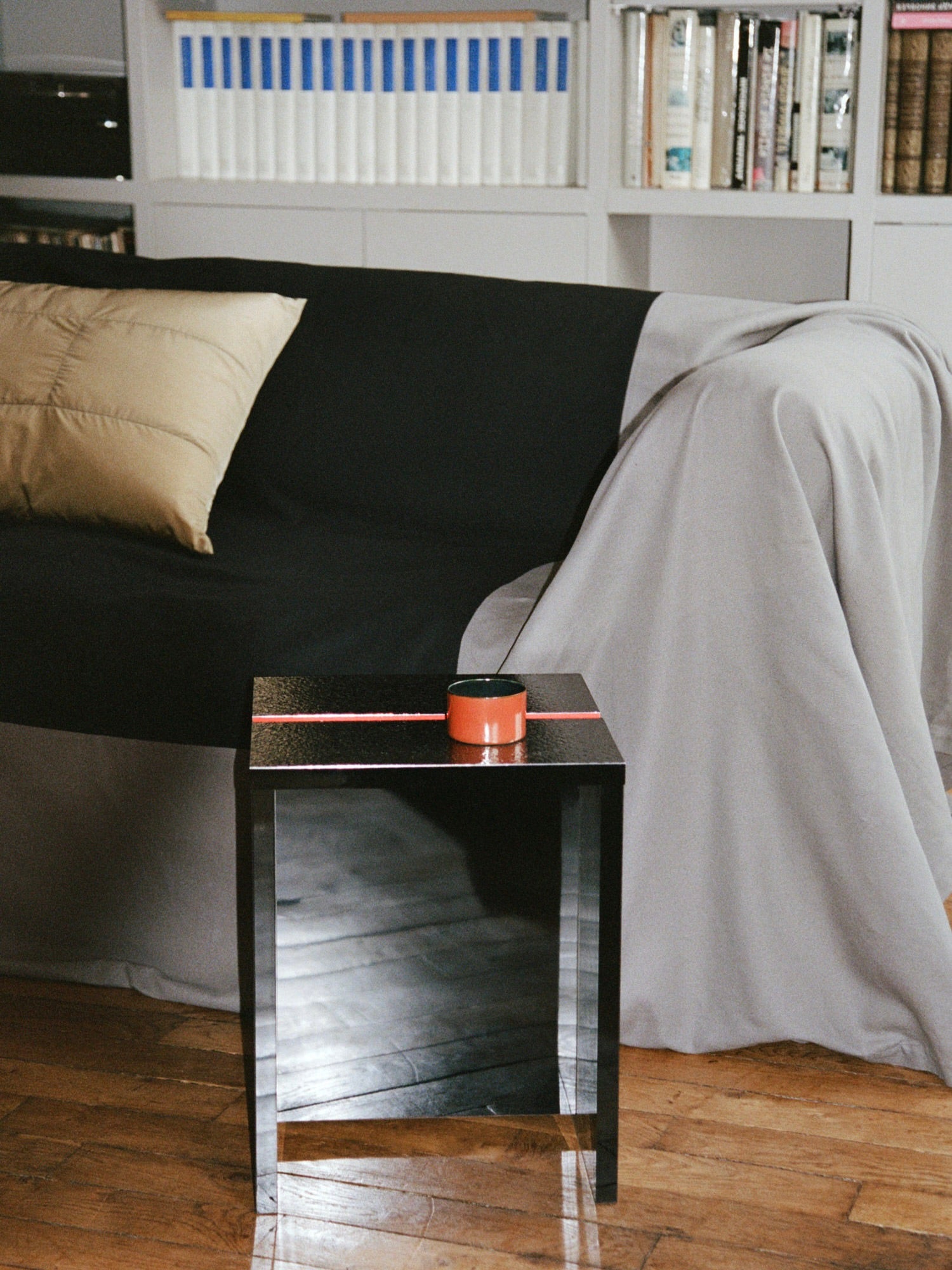 A cozy living room scene features a black and gray draped sofa, accented with a beige pillow, alongside the TEGET Mara Gap Stool in Wet Black. The stool holds an orange mug. A bookshelf showcases books on laminated furniture, while the wooden floor adds warmth to the space.