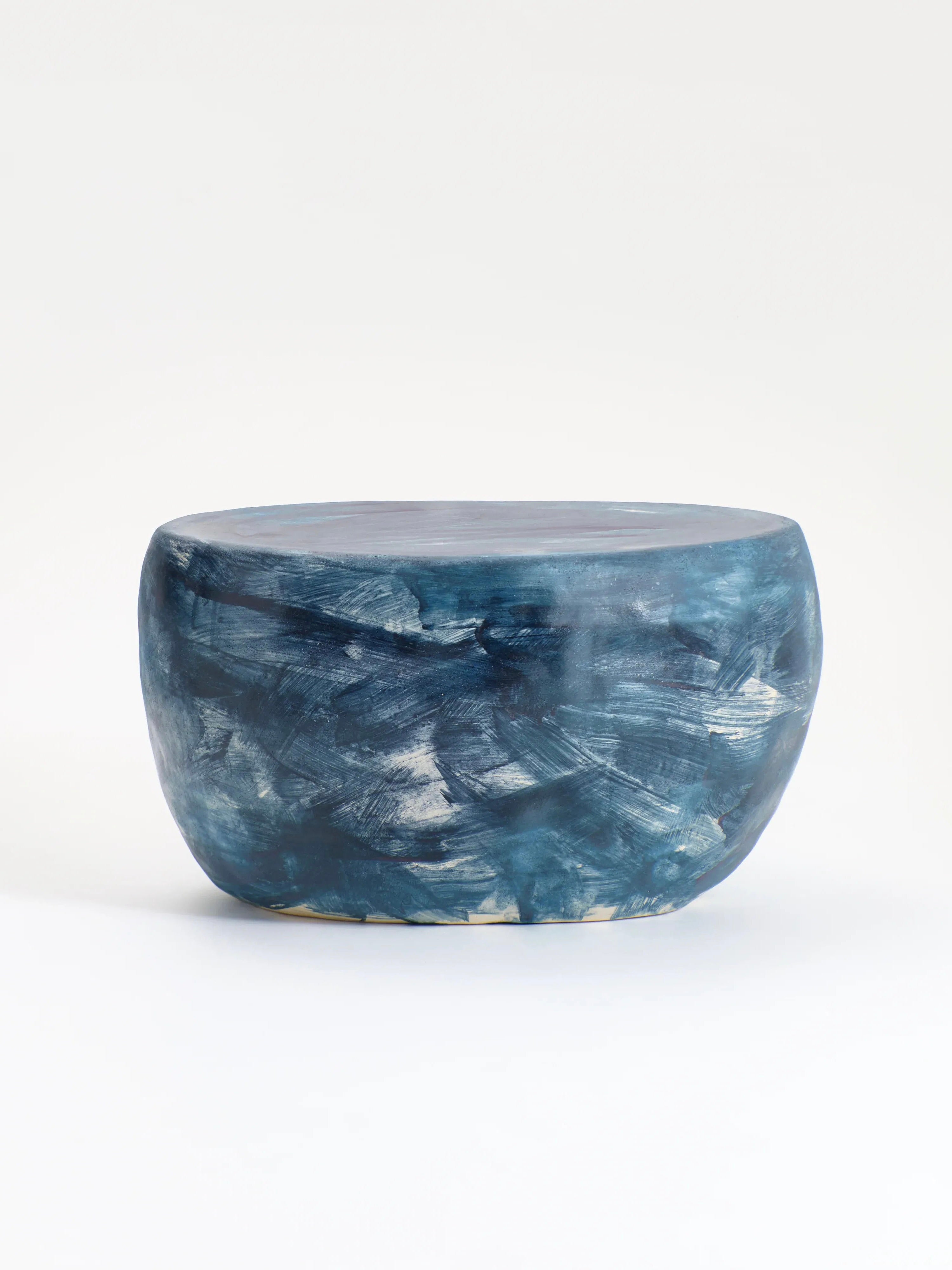 The Ceramic Coffee Table by Project 213A is a small, round, hand-sculpted piece with a flat top and cylindrical shape. It features swirling brush strokes in various shades of blue and hints of white glaze creating a marbled effect, all set against a plain white background.