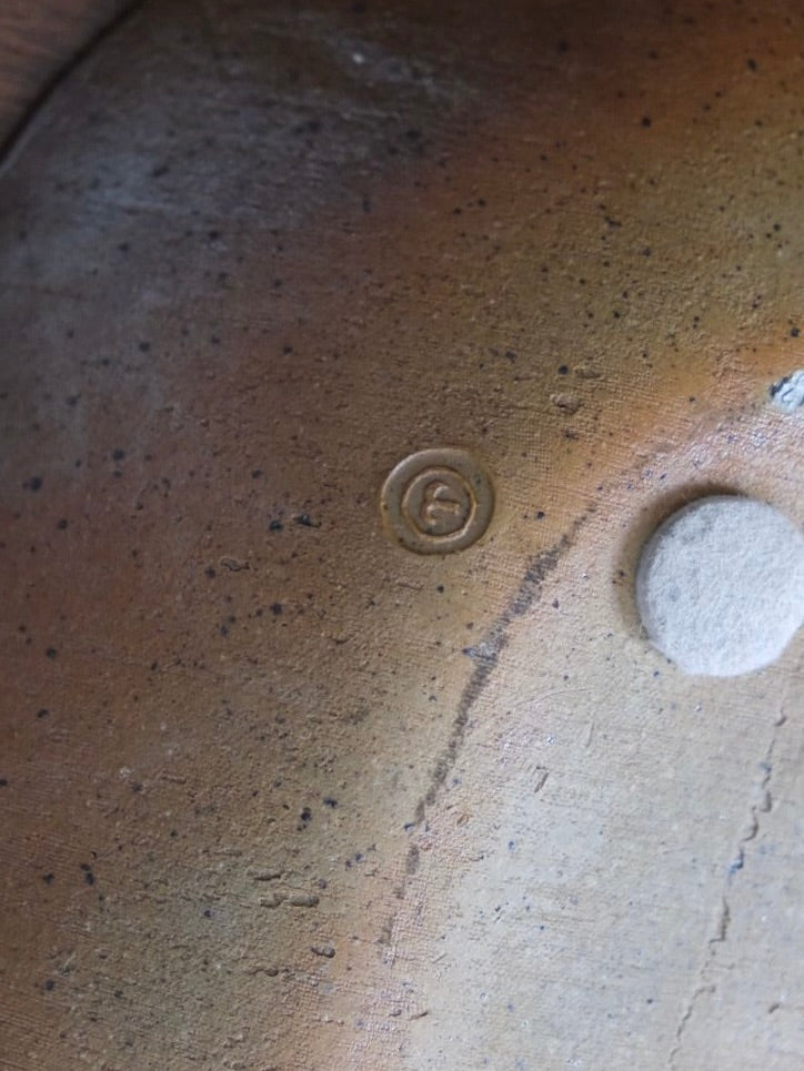 Product Description: A close-up of the Large Decorative Stoneware Dish by LA NOUVELLE GALERIE reveals a textured glazed surface, adorned with a small circular stamp embossed with a stylized "E" symbol. The speckled surface showcases a gradient of earthy tones, transitioning from light tan to dark brown, reminiscent of Pierre Culot's exquisite craftsmanship.