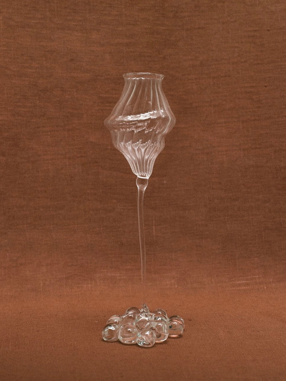 Against a brown fabric background, the uniquely shaped Orphée Liqueur Glass by Justine Menard stands out with its intricate design. This mouth-blown, handmade glassware features a wavy, bulbous form at the top, narrowing into a slender stem that culminates in a cluster of small glass spheres at the base. The set includes two exquisite pieces.