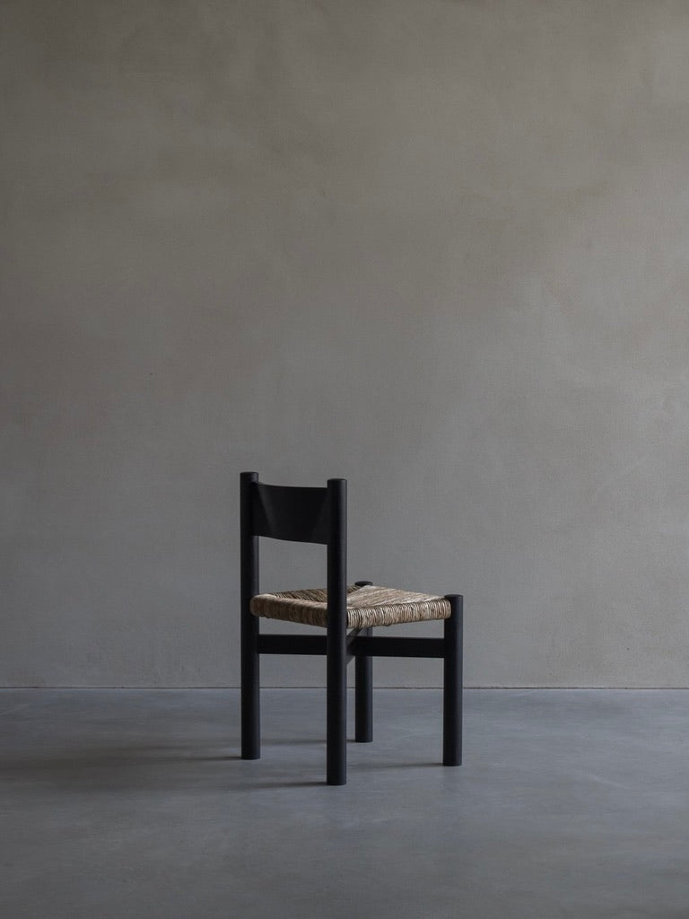 The 'Les Allues' straw chair by Bicci de' Medici, made from Mahogany wood with a woven seat, is placed sideways against a textured gray wall on a smooth concrete floor, highlighting its minimalist craftsmanship. The space radiates a calm, empty atmosphere.