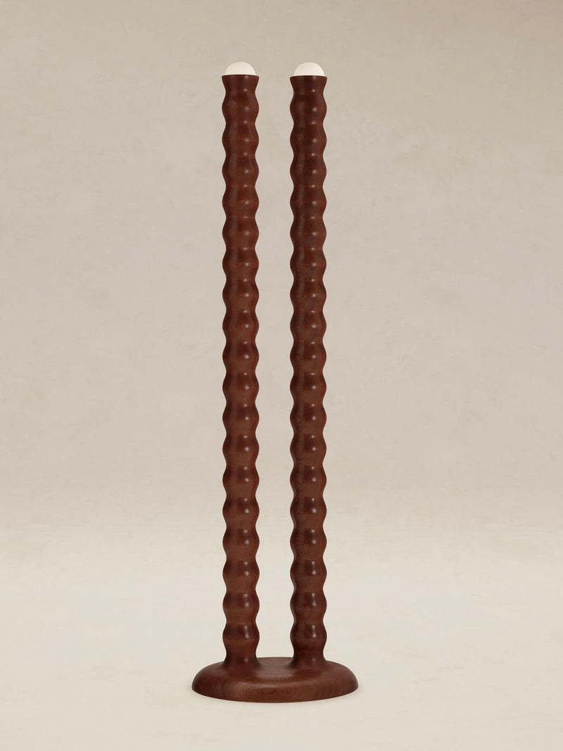 Introducing the "Tougarine" Floor Lamp by AÈDE Studios, featuring a pair of tall, wavy brown shafts crafted from cognac-red mahogany wood. These lamps elegantly stand upright on simple round bases with white tops, set against a neutral background.
