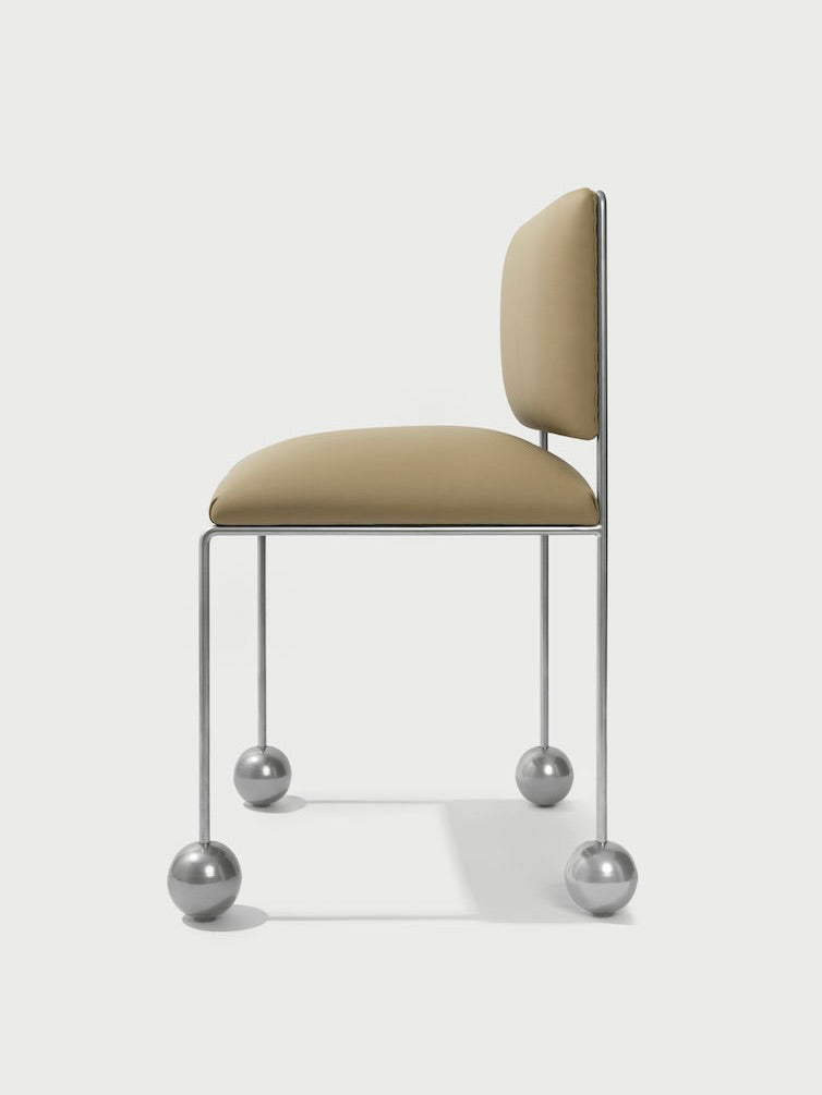 The Panorammma 'Ball Foot' Chair boasts a beige vegan textile cushioned seat and minimalist metallic frame, highlighted by its unique spherical metal feet, offering a modern design with sustainable luxury.