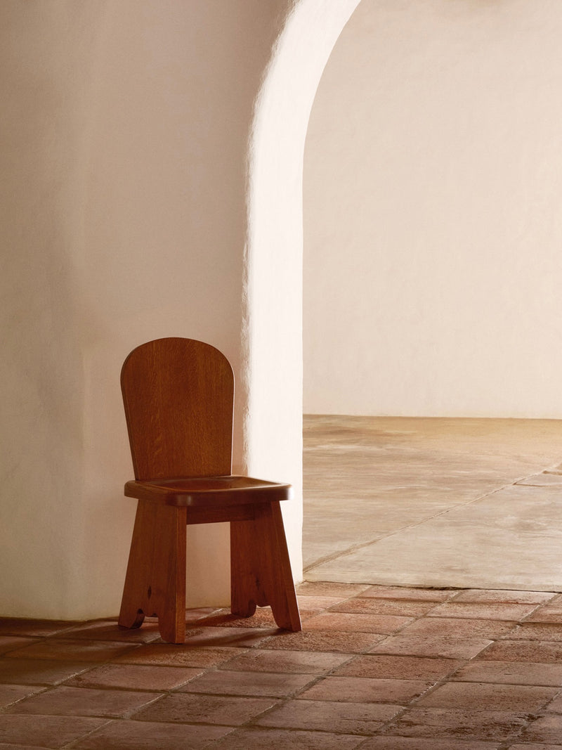 A Honey French Oak Wood Rambling Chair by Lemon sits against a white, textured archway wall on reddish-brown tiles. The warm minimalistic atmosphere with soft natural lighting suits contemporary living perfectly.