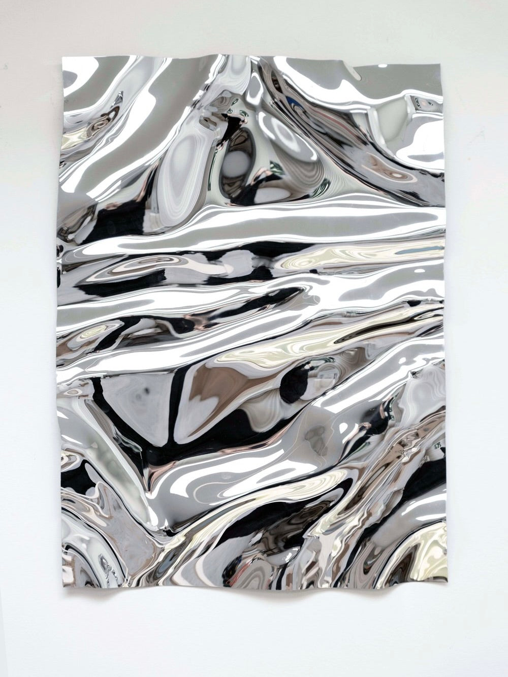 The Psychedelic Mirror by Caia Leifsdotter, a handcrafted masterpiece of crumpled, highly reflective metallic material, is elegantly hung against a plain, light-colored wall. The surface creates an abstract pattern of light and dark areas due to reflections and distortions, resembling a handmade sculptural mirror.