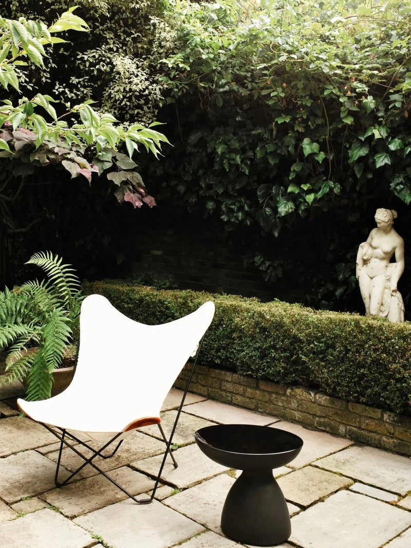 A garden setting features the "Cleo" Ceramic Side Table by Ransom & Dunn alongside a white butterfly chair on a stone patio. The area is enveloped in lush greenery, including ivy and ferns, while a classic statue peeks through dense foliage and neatly trimmed hedges.