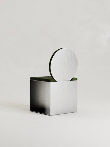 The 'Fragua' Chair by Siete Studio is a minimalist design with a geometric form—a metallic cube base with a disc partially inserted on top, showcasing metallurgical craftsmanship. It has a sleek, smooth finish set against an off-white background.