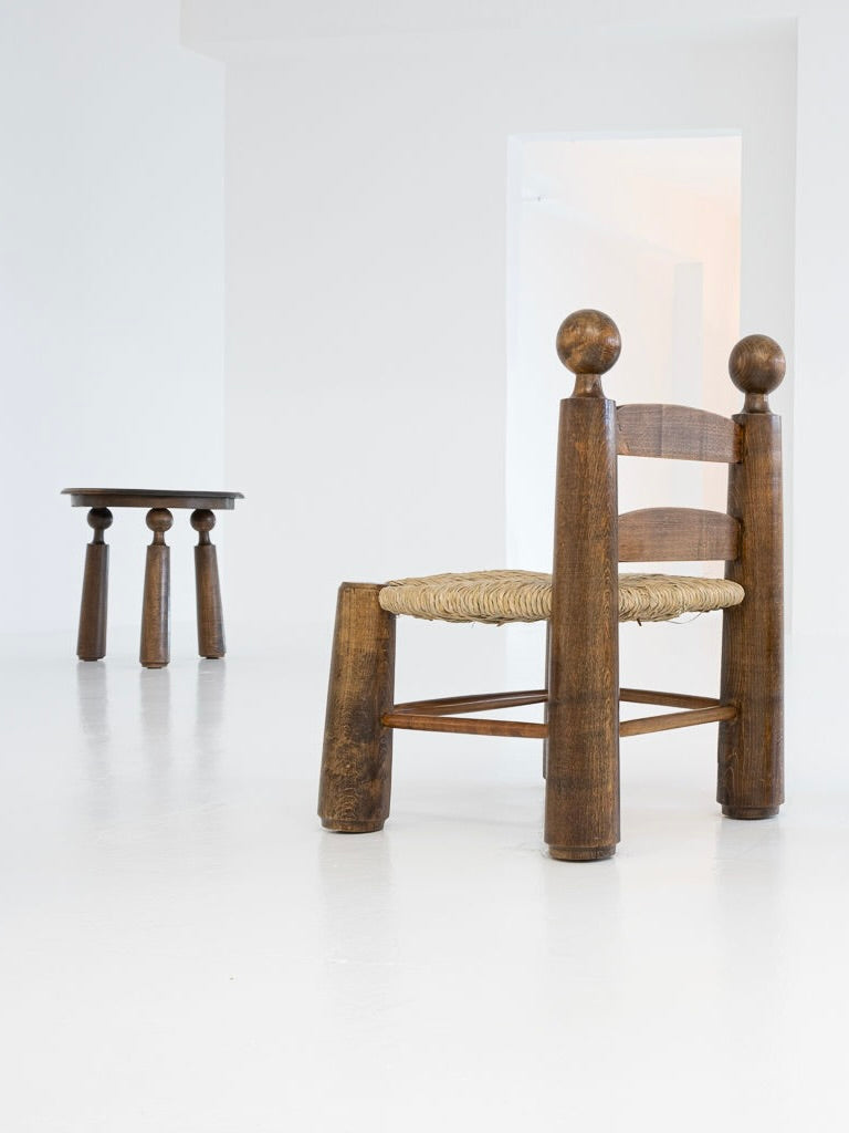 In the foreground is a large solid oak chair from the 1940s, crafted in Charles Dudoyut's style with thick cylindrical legs and a woven seat. Set against a white backdrop, a smaller round table accompanies it, all part of antibeige's set of two chairs and one table.