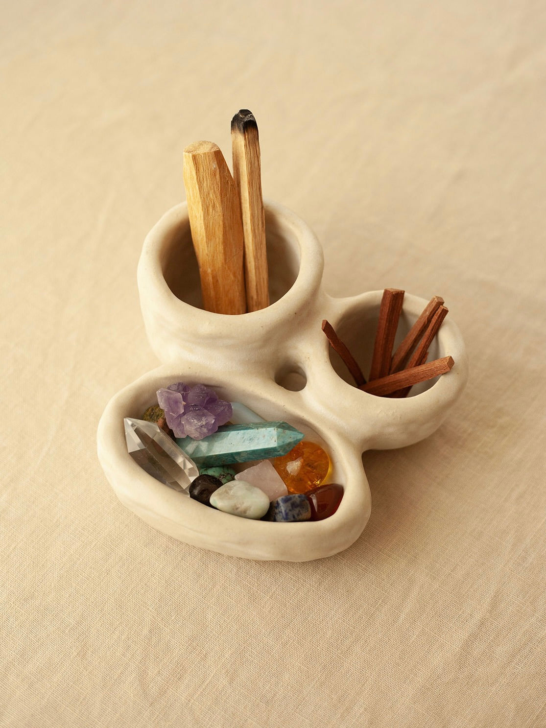 A cream-colored stoneware holder from INI Ceramique's 'Organic Fusion N°3' collection contains various crystals, palo santo sticks, and cinnamon sticks displayed on a light tan fabric background. The colorful crystals vary in shape and size, adding vibrancy to the scene.