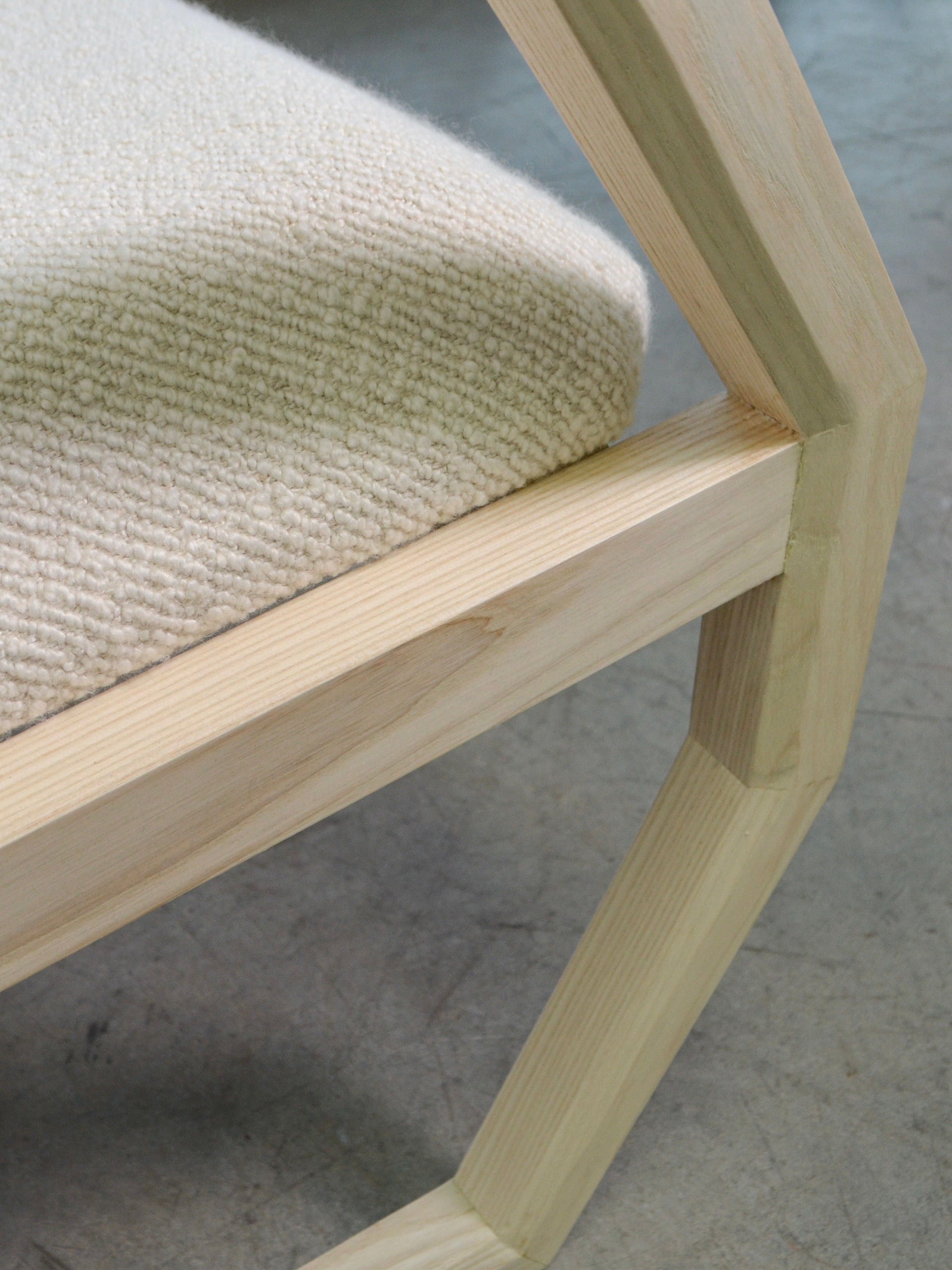 A close-up of the Marcel Accent Chair by Nathan Chintala Design LLC showcases its geometric wooden frame and cushioned seat upholstered in a textured, cream-colored fabric. The angled joint of the chair's leg is visible on a smooth concrete floor. This piece is perfect for those seeking both style and comfort.