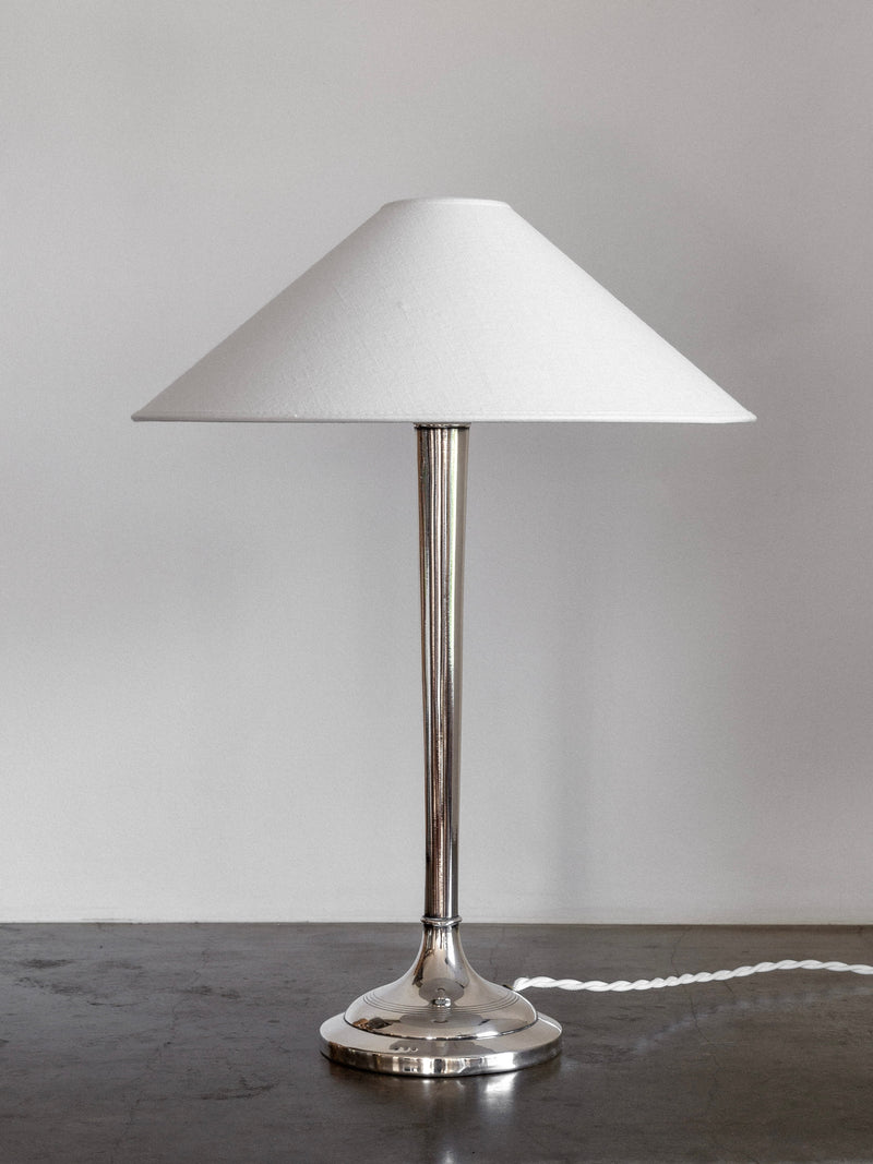 An elegant Spigel Silver-Plated Table Lamp from the early 1900s boasts a sleek, reflective base paired with a conical white lampshade. The lamp, showcasing subtle Art Deco style, rests on a dark gray surface with its power cord visible against a plain light gray background.