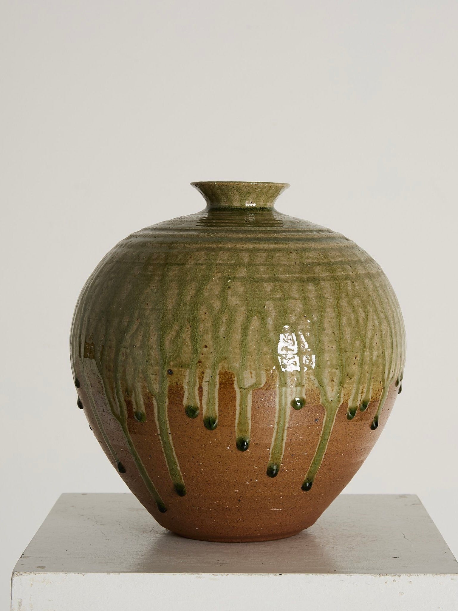 The Drip Glazed Vase by Ancien et Jolie is a mid-century style ceramic piece featuring a brown base and glossy green drip glaze inspired by Hokkaido, Japan, elegantly displayed on a white pedestal against a simple backdrop.