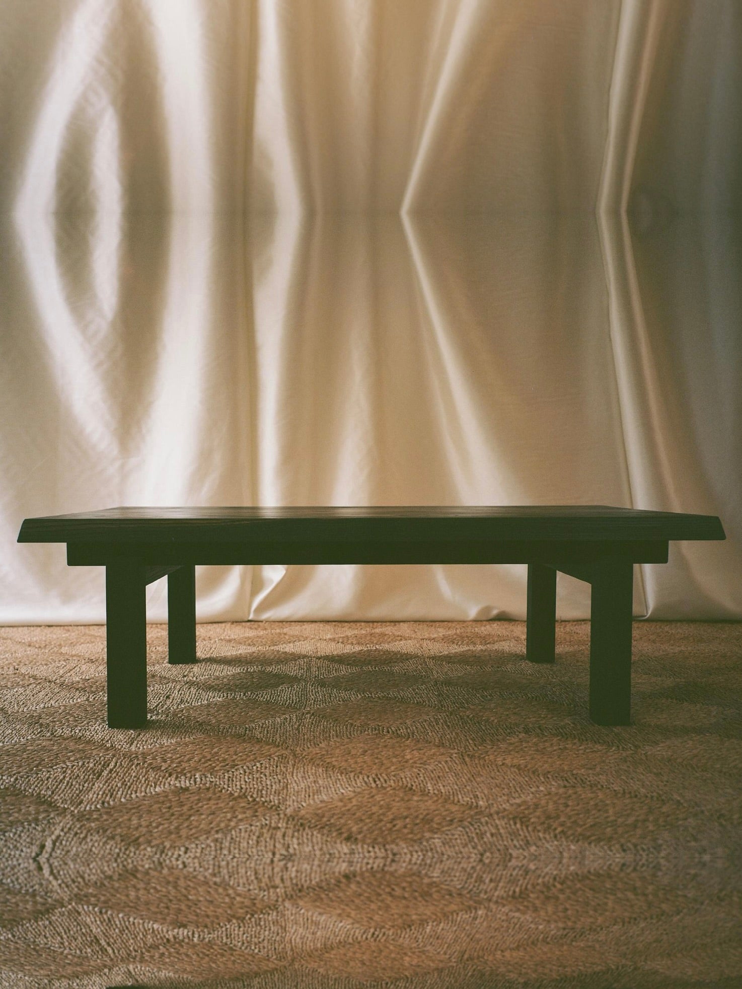 The SWM STUDIO Lantern Coffee Table, made of solid oak with four legs, stands elegantly on a textured rug. Softly draped illuminated curtains in the background evoke Japanese aesthetics, creating a warm and cozy atmosphere.