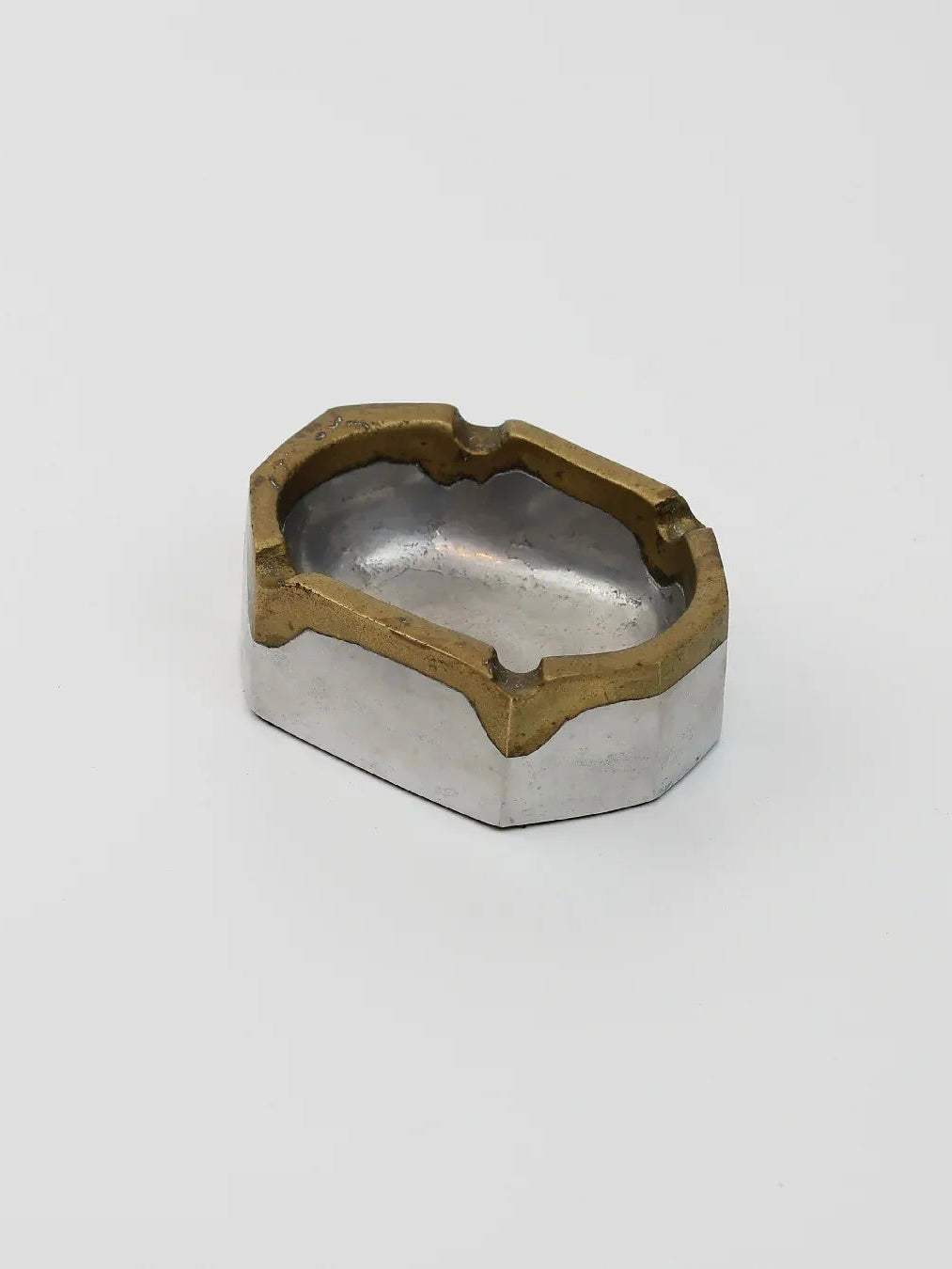 Product Description: The Brutalist Ashtray by Boga Avante Shop is a small, hollow hexagonal object made from brass and steel. Featuring a silver base and gold-toned top edges, its design is reminiscent of David Marshall's brutalism. It has an indented, curved center against a white background.