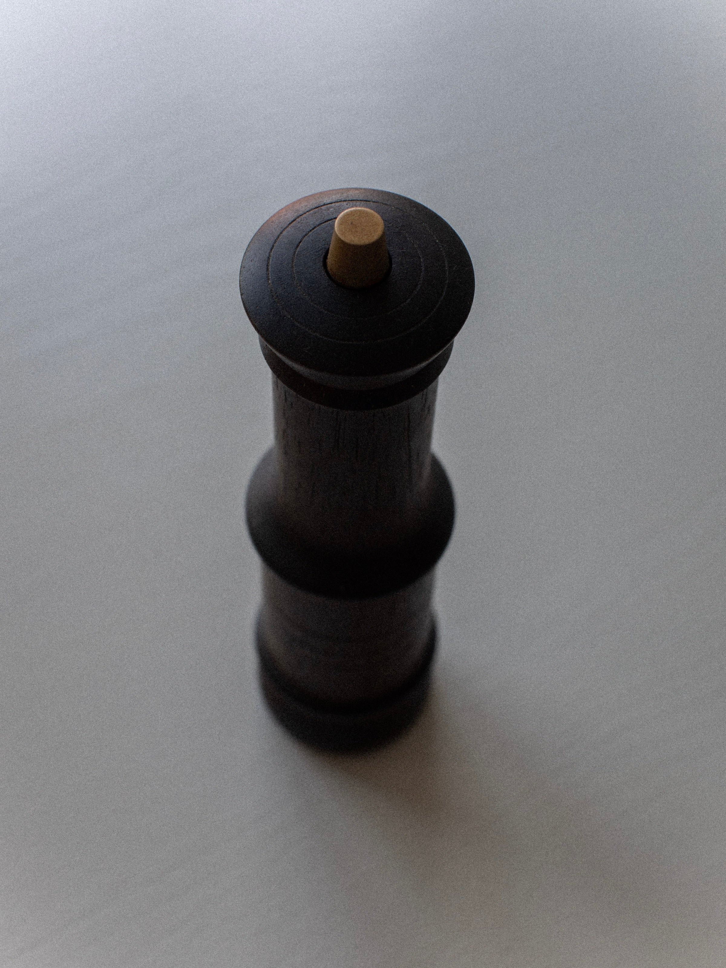 Image of the Out For Lunch Wenge Wood Grinder standing upright on a smooth, light-colored surface. The grinder, crafted from wenge wood, is cylindrical with a rounded top and a knob at the center for grinding. The lighting casts subtle shadows, enhancing its slightly vintage appearance reminiscent of Denmark's elegant design aesthetics.