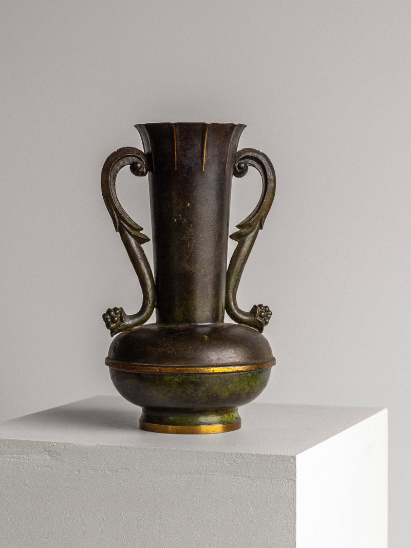An Art Deco Bronze Vase by Spigel, featuring a dark patinated finish, elaborate handles resembling stylized dragon heads, and a narrow neck leading to a rounded base reminiscent of a neoclassical urn shape, is displayed on a white pedestal against a plain gray background.