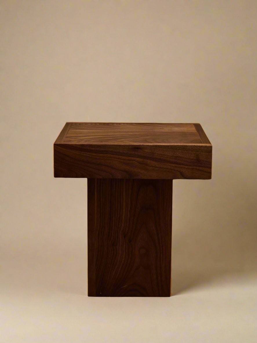 The Jura Pedestal, a handcrafted piece by Ah Um Design Studio, features a square top and rectangular base. Positioned on a beige floor with beige curtains in the background, it is masterfully created from walnut wood. The elegant side table boasts a smooth, polished surface that beautifully showcases the natural grain, making it an exquisite piece of display furniture.