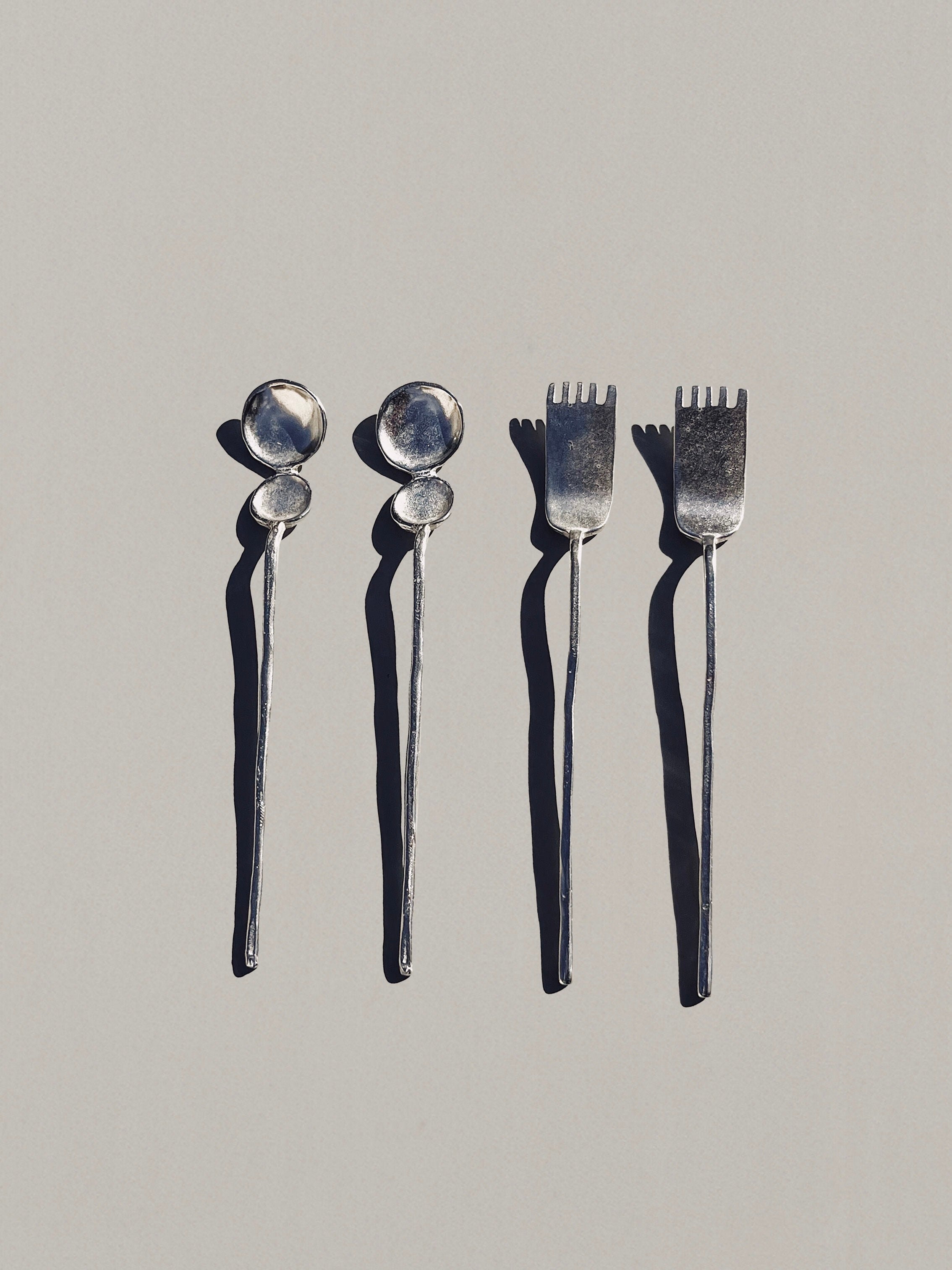 A minimalist image features the "Artifact" Tea Cutlery by Hors-d'Oeuvre, with two silver spoons on the left and two forks on the right, each casting long shadows on a light gray background. This handcrafted set displays a simple, elegant design that perfectly captures the essence of a refined dining ritual.