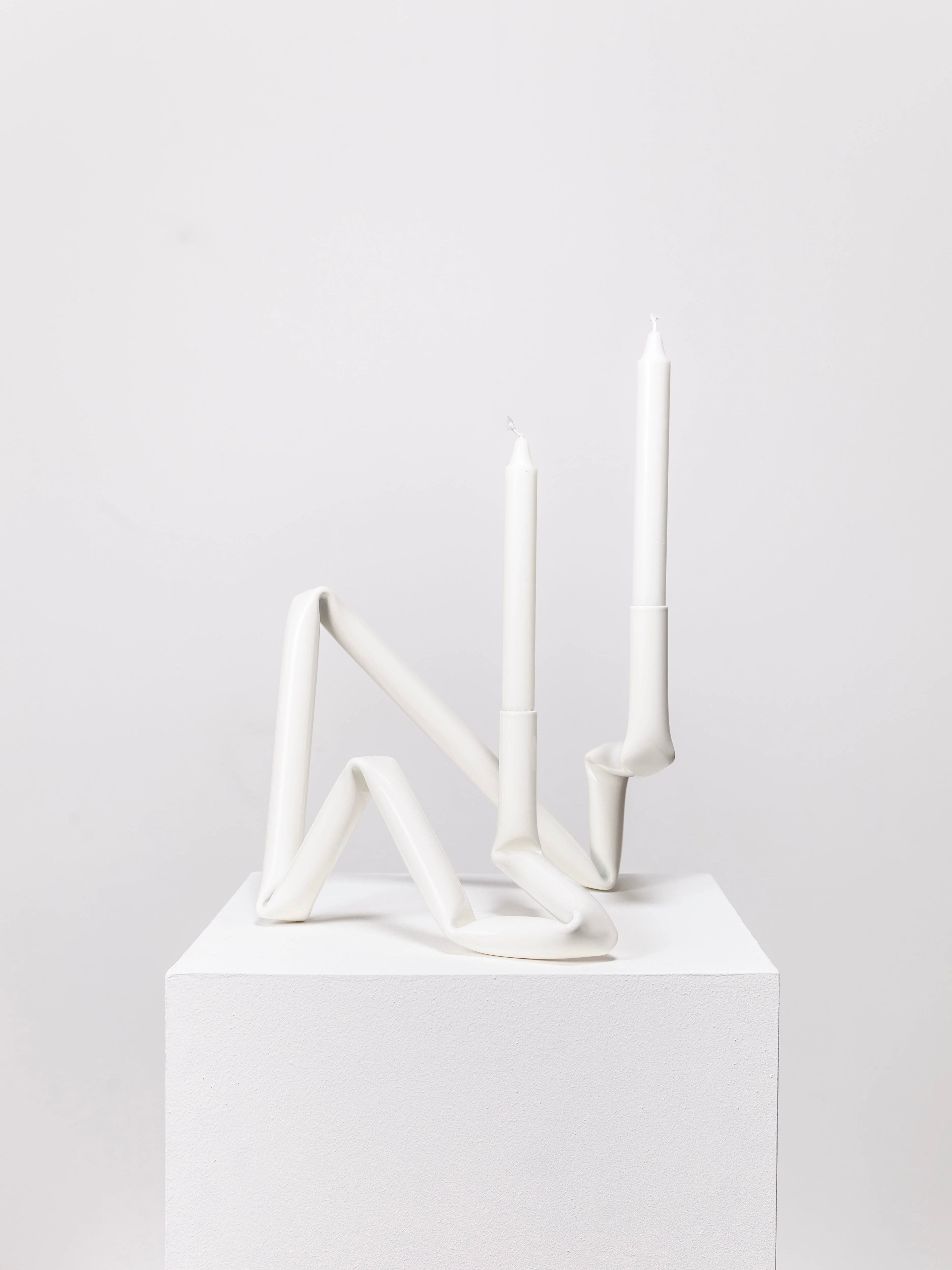 The Bucatini candle holder (White) from AOT STUDIO's Primi collection boasts a unique, fluid geometric design. Crafted from aluminum tubing, it holds two tall, unlit white candles. The minimalist structure appears twisted and modern, sitting on a plain white pedestal against a plain white background.
