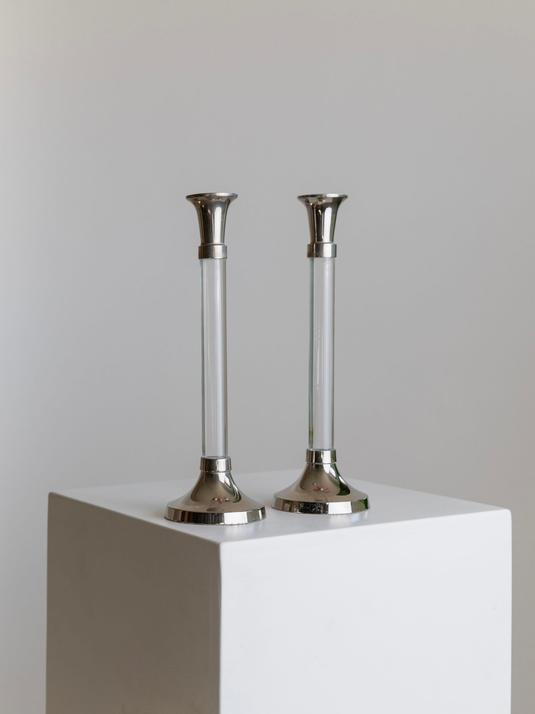 A pair from the Bottega Jacobs' Set of 2 Candleholders, featuring silver and glass construction reminiscent of vintage candlesticks, are elegantly positioned side by side on a white pedestal against a neutral backdrop. The candleholders boast a sleek, reflective finish that adds a modern and sophisticated touch to the display.