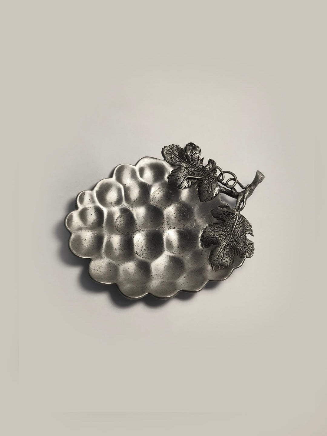 This decorative piece, the Grape Vide Poches by Les Objoies, showcases a grapevine motif with embossed grape leaves and vines. Crafted from pewter, it is shaped like a grape cluster and elegantly rests against a light gray background.