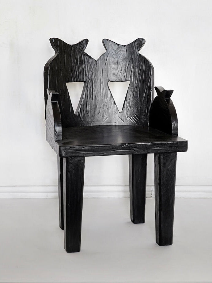 The Pomegranate Chair by Rooms Studio is a black wooden chair made from reclaimed oak, featuring a textured surface and a backrest with two stylish cat silhouettes. The armrests resemble small cats, highlighting rustic craftsmanship, all set against a plain white background.