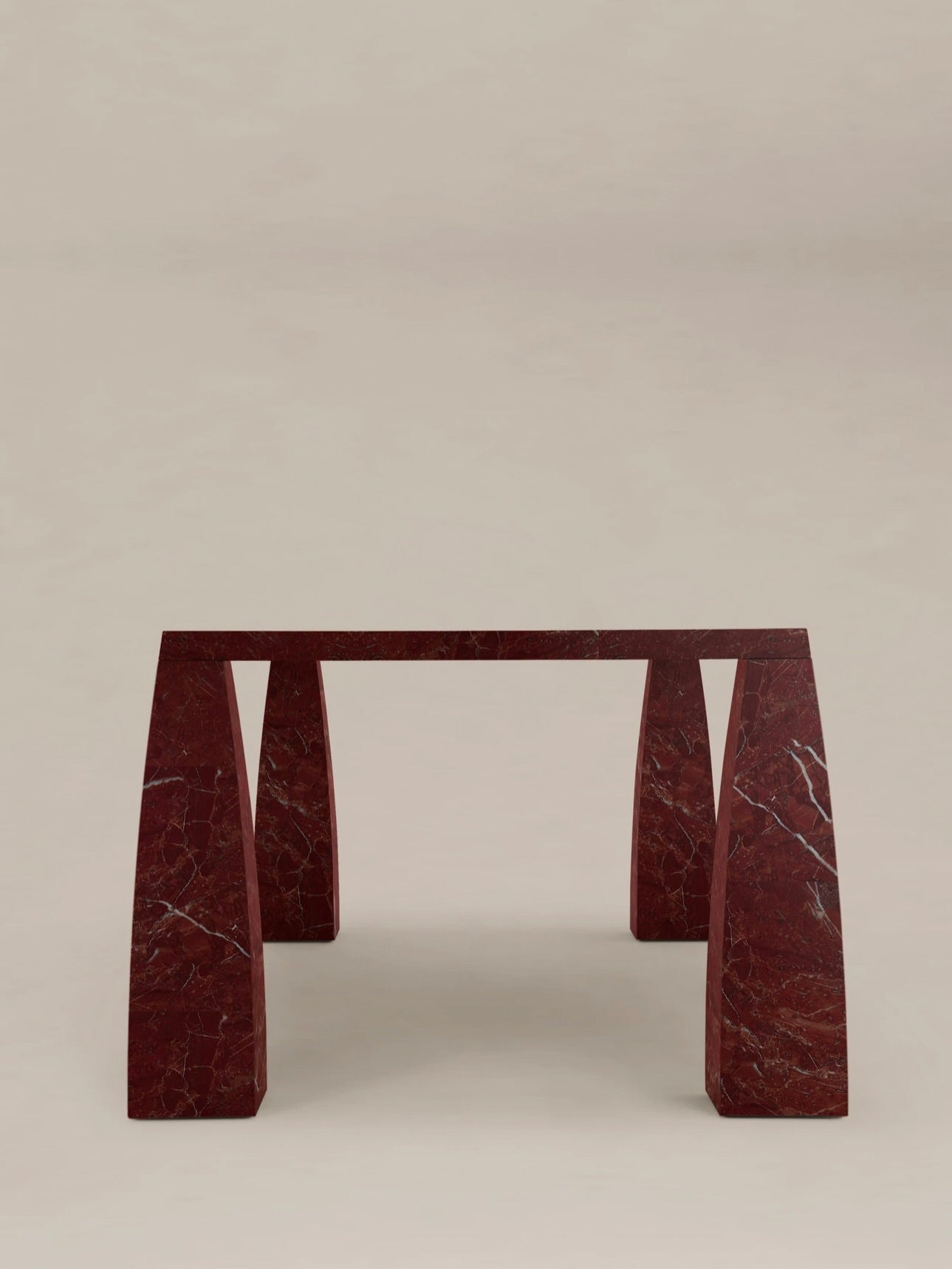 The "Khram" Dining Table by AÈDE Studios boasts a striking design reminiscent of medieval Russian architecture, featuring two substantial angled legs that support its flat, rectangular top. This red marble piece, with natural white veining, is elegantly modern and stands out beautifully against a simple, neutral background.