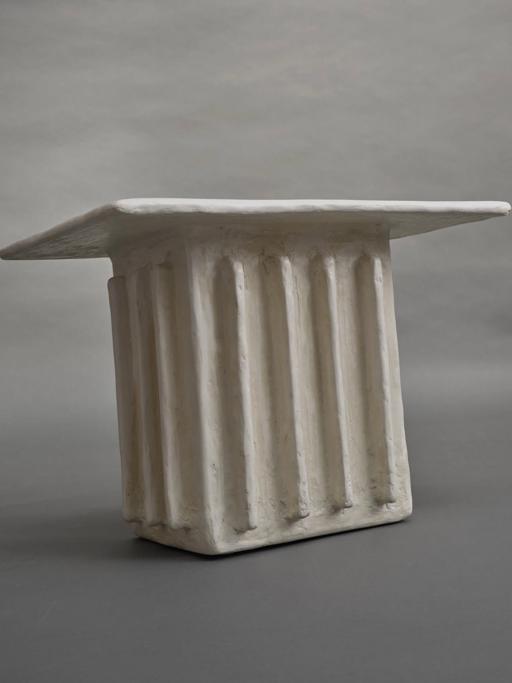 Set against a plain grey background, the Columnar Side Table by Ombia Studio features a sculptural design made from raw white plaster, resembling a rectangular pedestal with fluted sides and a flat top, crafted in a textured stone-like material.