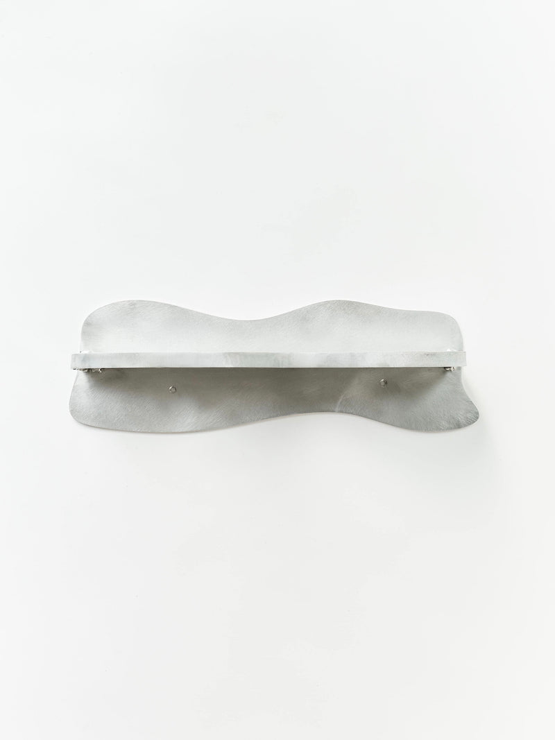 The Middle Shelf by Six Dots Design is a versatile, minimalist bathroom shelf with a wavy design and smooth metallic surface, offering a sleek and modern appearance that elegantly contrasts against a plain white background.