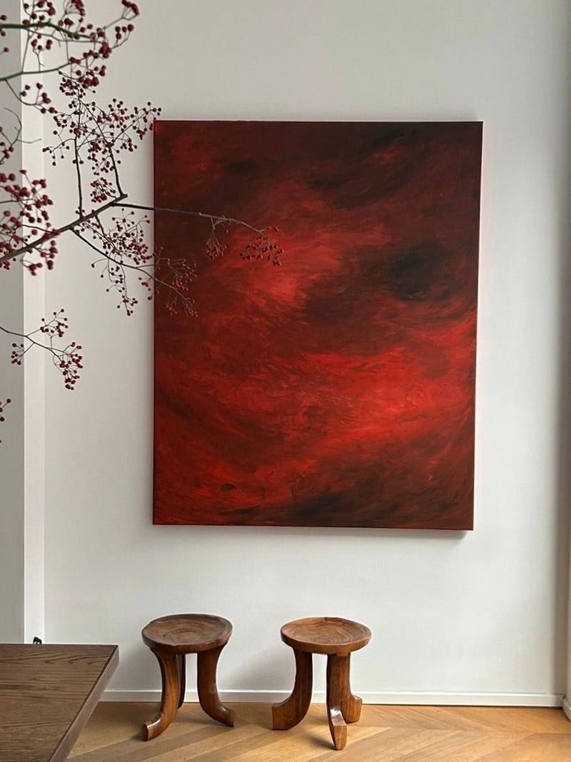 A large abstract piece titled "Ember’s Whisper" from Mattias Storm, with swirling red and dark tones, is displayed on a white wall. It is elegantly framed in oak or walnut. Beneath it sit two small wooden stools, while a branch with small leaves extends from the left side of the image.
