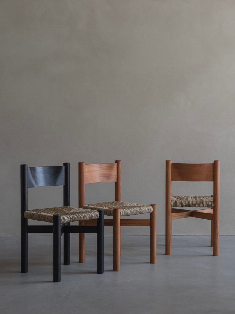 Three 'Les Allues' chairs by Bicci de' Medici, expertly crafted from Mahogany with straw weaving, stand side by side. Two feature natural wood tones, while one is black. These chairs highlight fine craftsmanship and simple yet sturdy designs against a neutral backdrop.