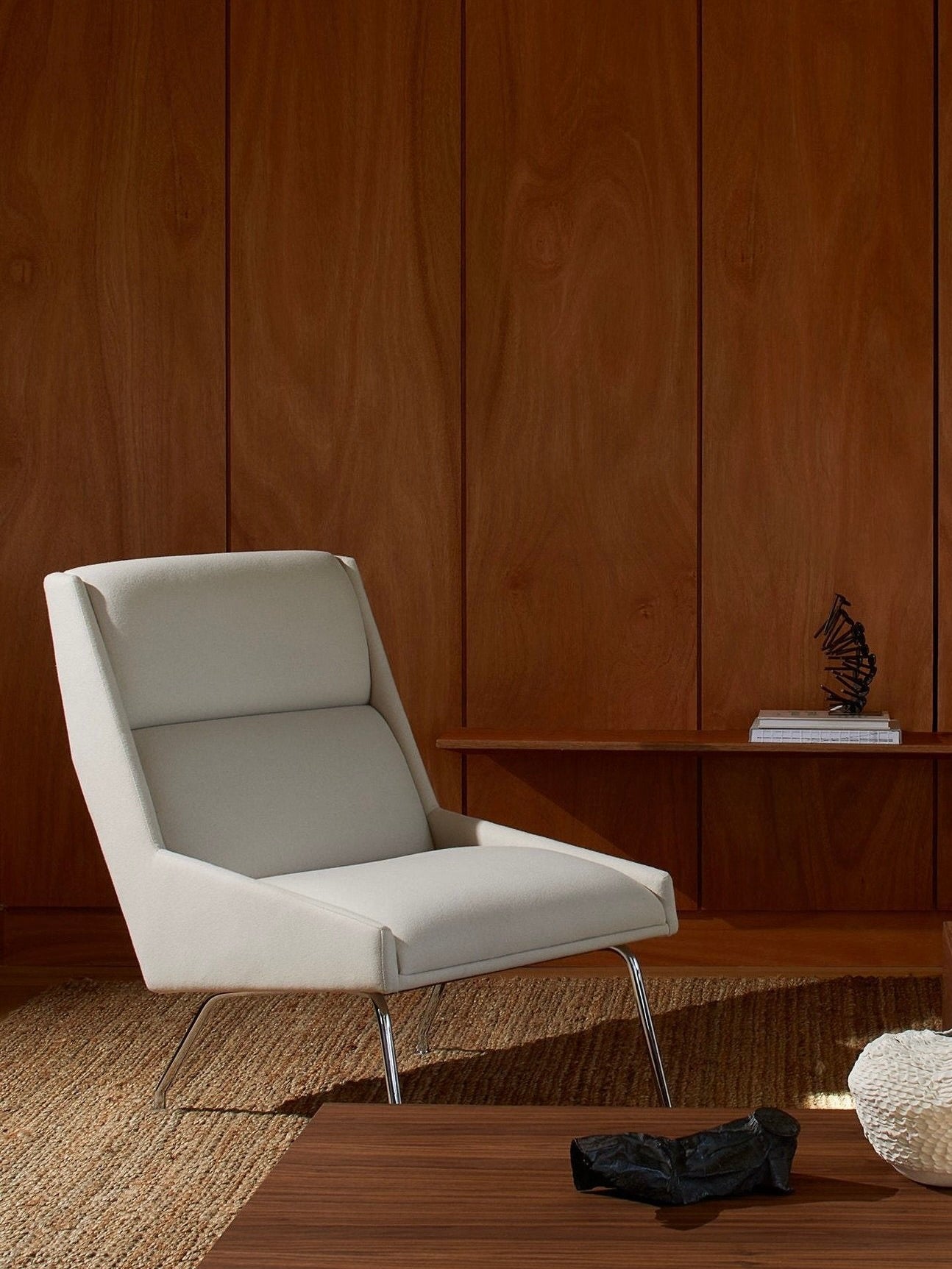 A modern room showcases the "Kent" Armchair by Lemon in a light gray finish, echoing Italian 1950s design, positioned on a woven rug. The background is adorned with wooden wall panels and a low wooden shelf displaying a small sculpture and books, while a wooden table is partially visible in the foreground.