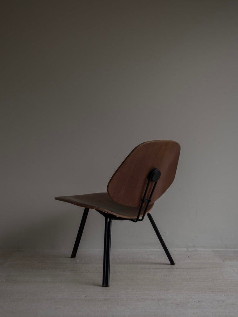 A minimalist wooden chair, inspired by Italian Mid-Century style, featuring a curved backrest and black metal legs, is placed against a gray wall on a light wood floor. Angled to the side, its sleek modern design mirrors Bicci de' Medici's Osvaldo Borsani - P31 chairs.