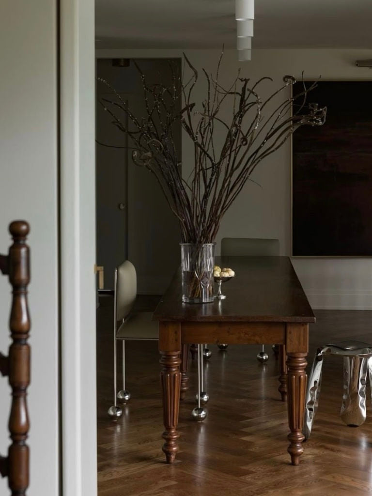 A dining room features a dark wooden table with a glass vase of tall, bare branches as the centerpiece. The minimalist 'Ball Foot' chairs by Panorammma, crafted from vegan textiles with metal legs, rest on a polished wooden floor under subdued lighting, creating a modern rustic ambiance.