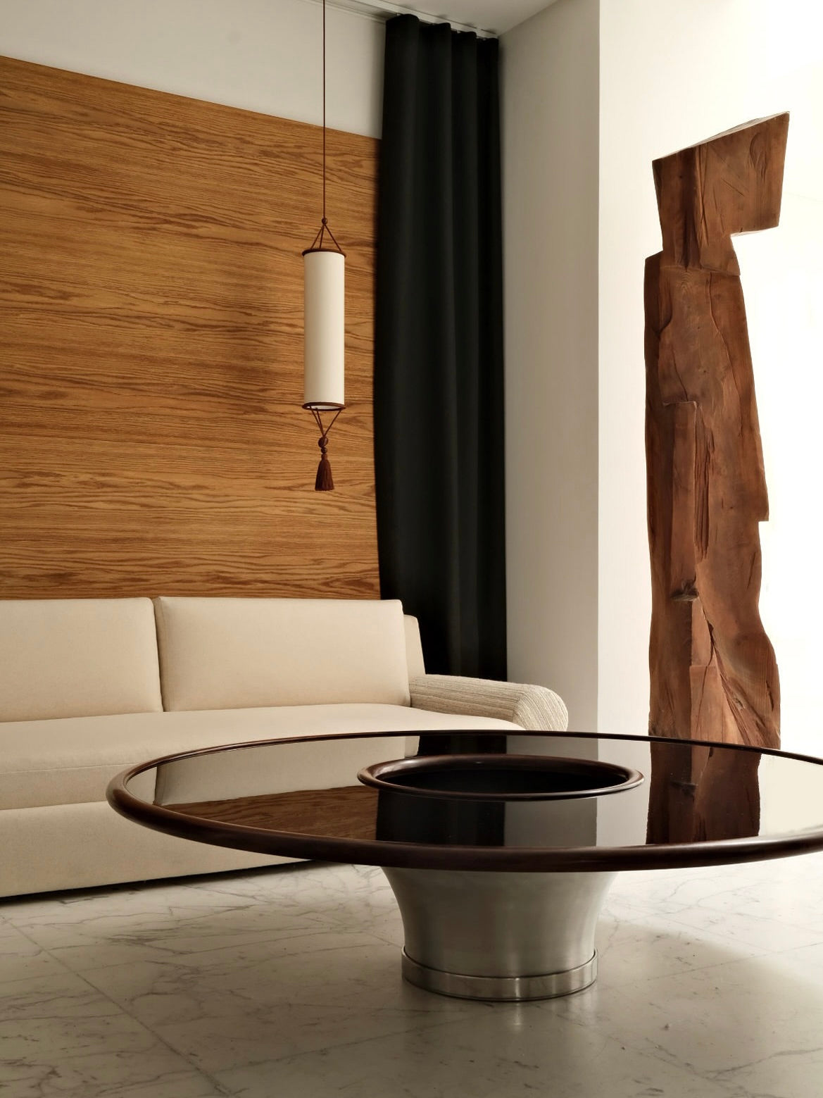 In a minimalist room, a beige sofa is set against a wooden accent wall, complemented by a cylindrical pendant light and a distinctive wooden sculpture. The "Loulou" Coffee Table by HAUVETTE & MADANI, featuring an embossed aluminum base and round glass top, rests on the marble floor. Dark curtains frame the bright window.