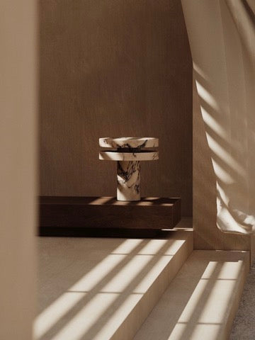 In a minimalist room, the 'M_014' Marble Table Lamp by Monolith rests on a wooden platform. Sunlight filters through blinds, casting shadows on cream walls and floor, enhancing the serene and elegant ambiance.