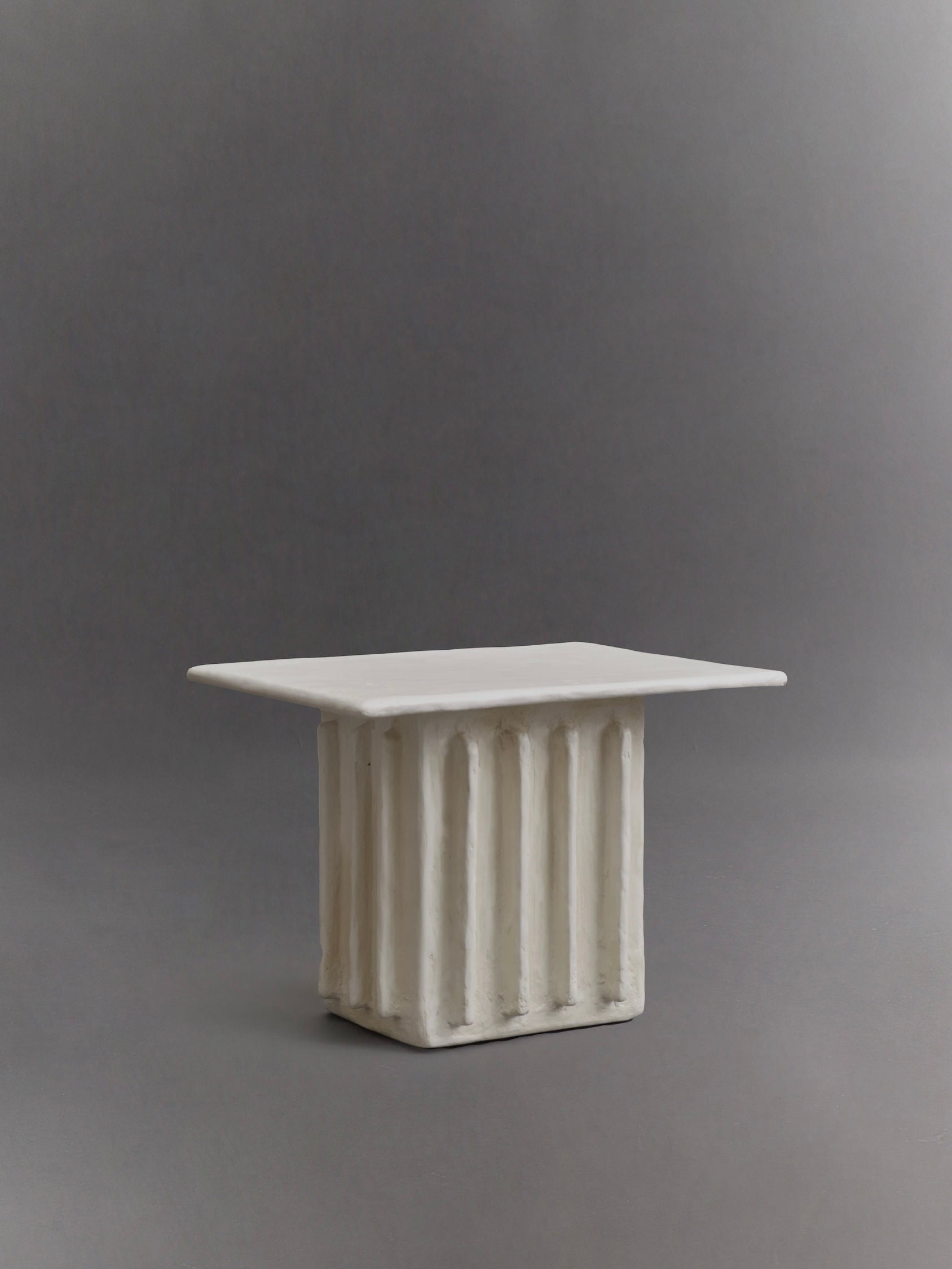 The Columnar Side Table by Ombia Studio is a rectangular, sculpted piece made from raw white plaster with vertical grooves, resembling a small table or pedestal, showcased against a neutral gray background.