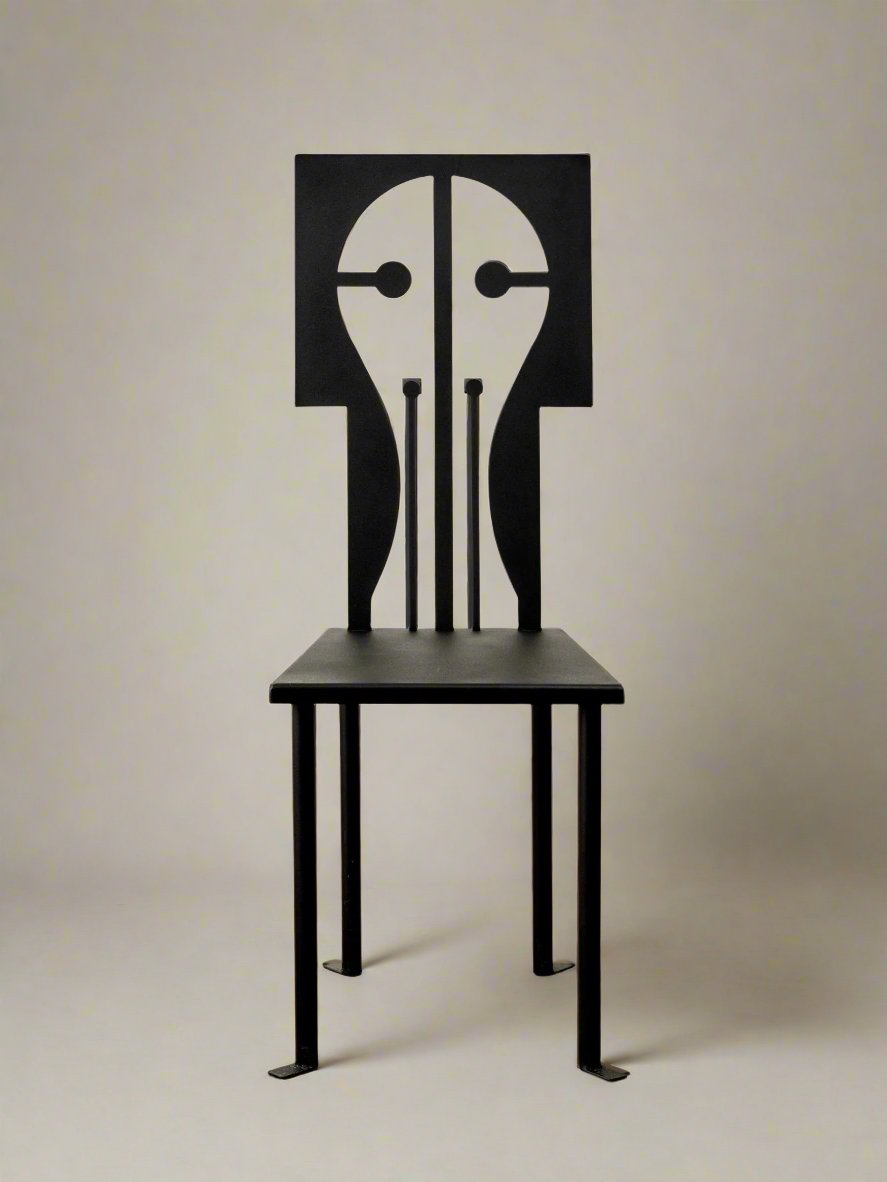 Introducing the Epona Chair in Iron and Black Powder Coat by Animate Objects. As part of the Rèveuse collection, this exquisitely handcrafted piece features a black frame with a distinctive geometric backrest reminiscent of a pen nib. It is pictured elegantly standing on a polished wooden floor against a simple white background.