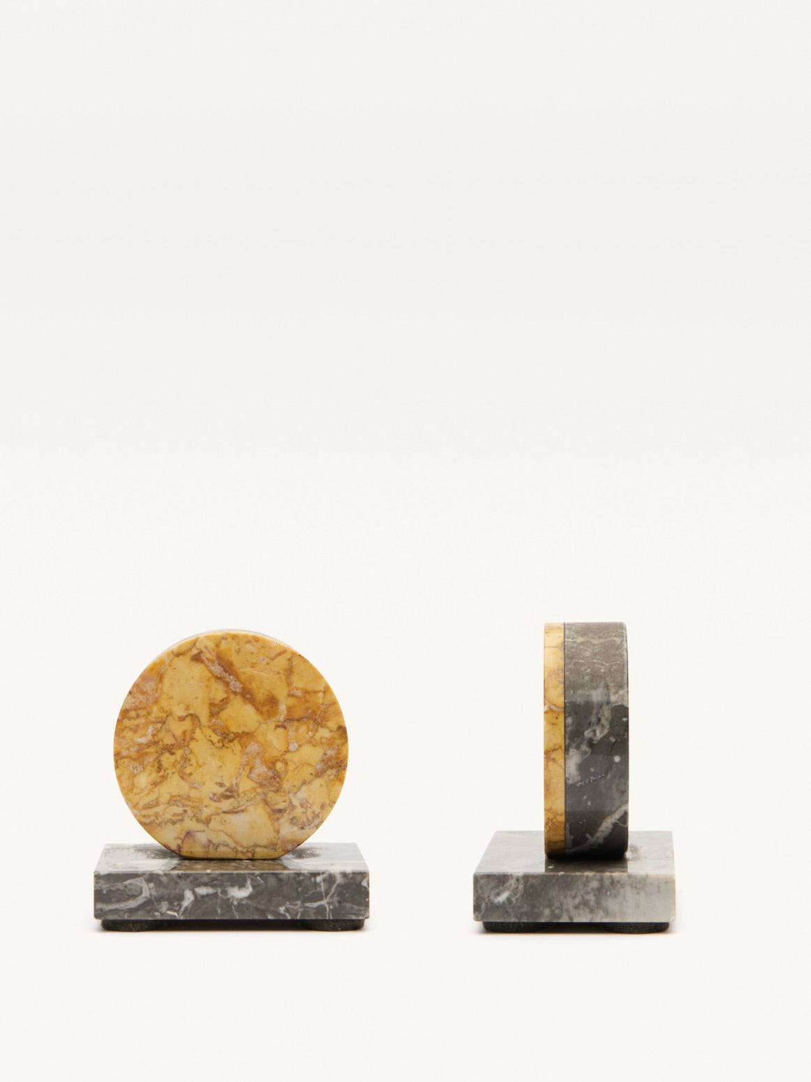 The Pair of Art Deco Marble Bookends by Galerie MLS showcases 1930s-inspired design with decorative circular stone tops and marbled patterns. Each circle sits on a square black marble base, displaying the full face on the left and a side view on the right.