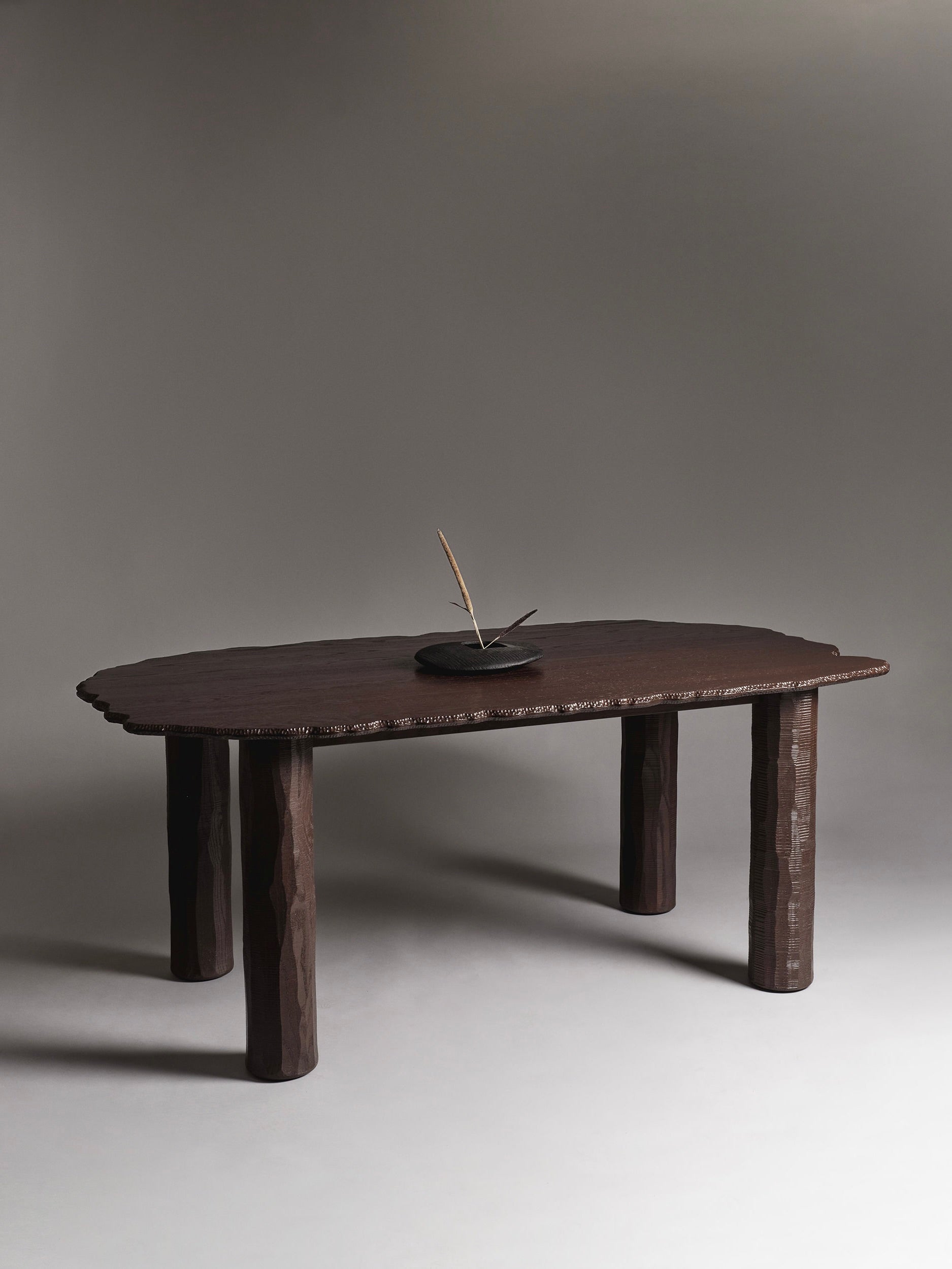 The Mare Dining Table by Ombia Studio is a large, dark hand-carved wooden table featuring a textured surface and four thick legs. Set against a plain gray background, it includes a minimalist sculpture and offers custom sizing to suit its functional yet elegant design.