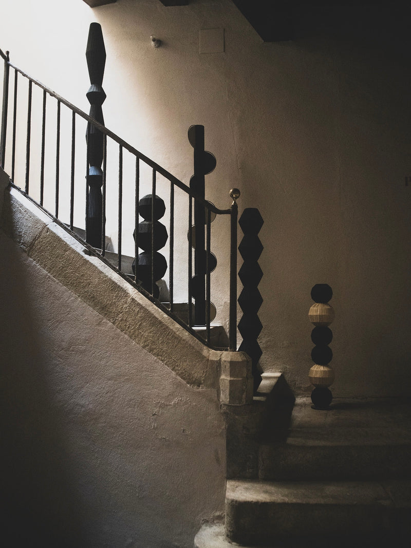 A dimly lit stone staircase with a black metal railing featuring large, round geometric balusters, ending in a post adorned with black and beige spheres, showcases the "Column V" Sculpture by ELIURPI, inspired by handmade Barcelona woven art.